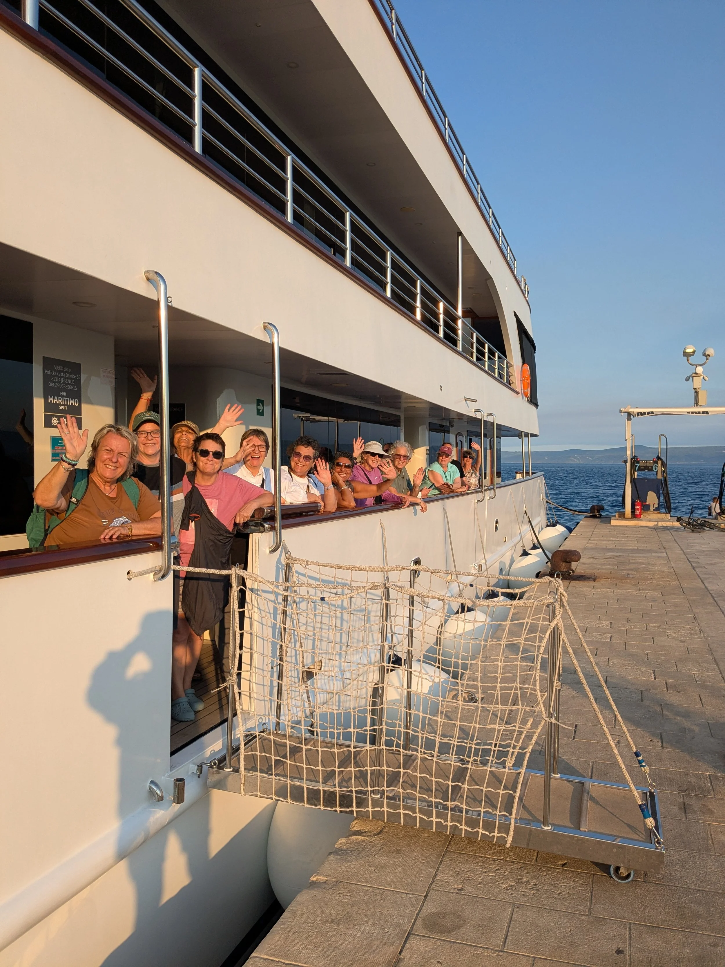 A group of women waving and smiling from a small white tender boat on the sparkling blue Adriatic sea. A crew member steers the boat, and the green Croatian coastline is visible in the background.