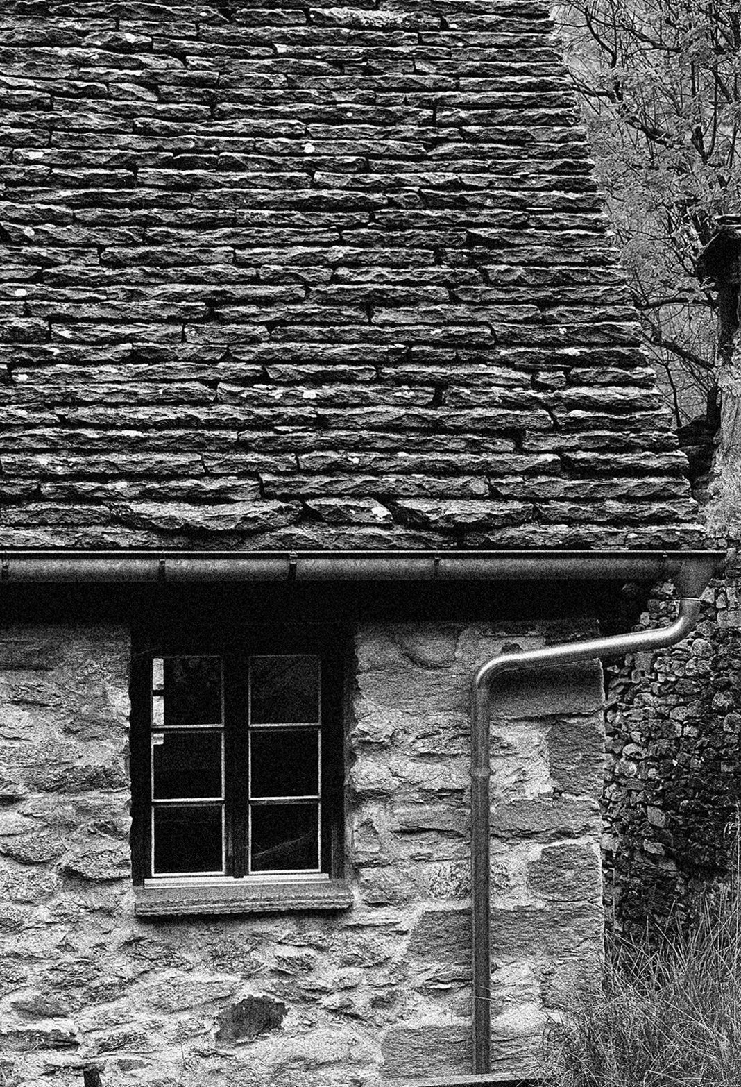 Black and white photo of a stone cottage wall with a small window and a metal drainpipe.