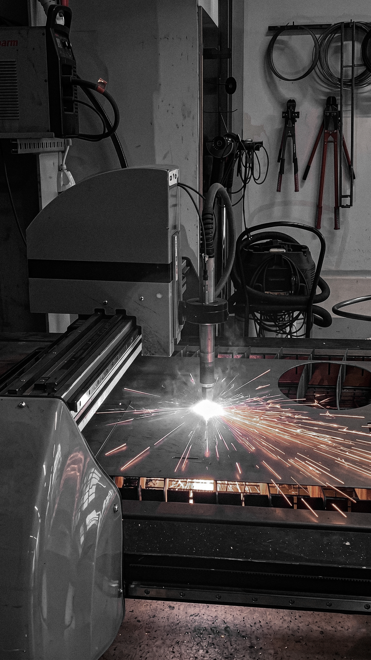 A welding machine is cutting or welding a metal surface, producing bright sparks, in a workshop with tools hanging on the wall.