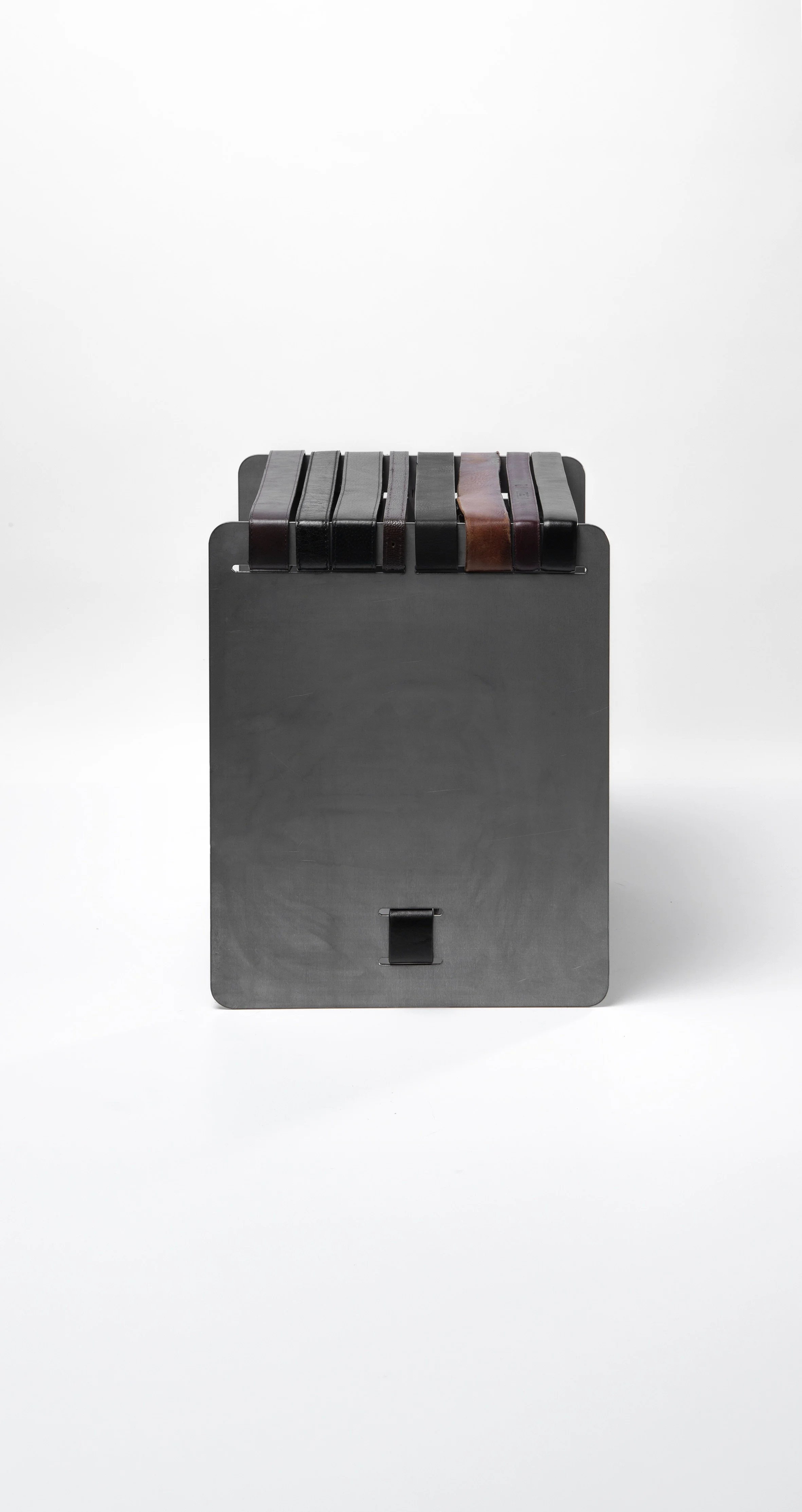 A black metal stool holding multiple leather belts in various shades of black, brown, and maroon, set against a plain white background.  Architecture and Design by Auliseo (Aurèle Eliseo Stauffer).