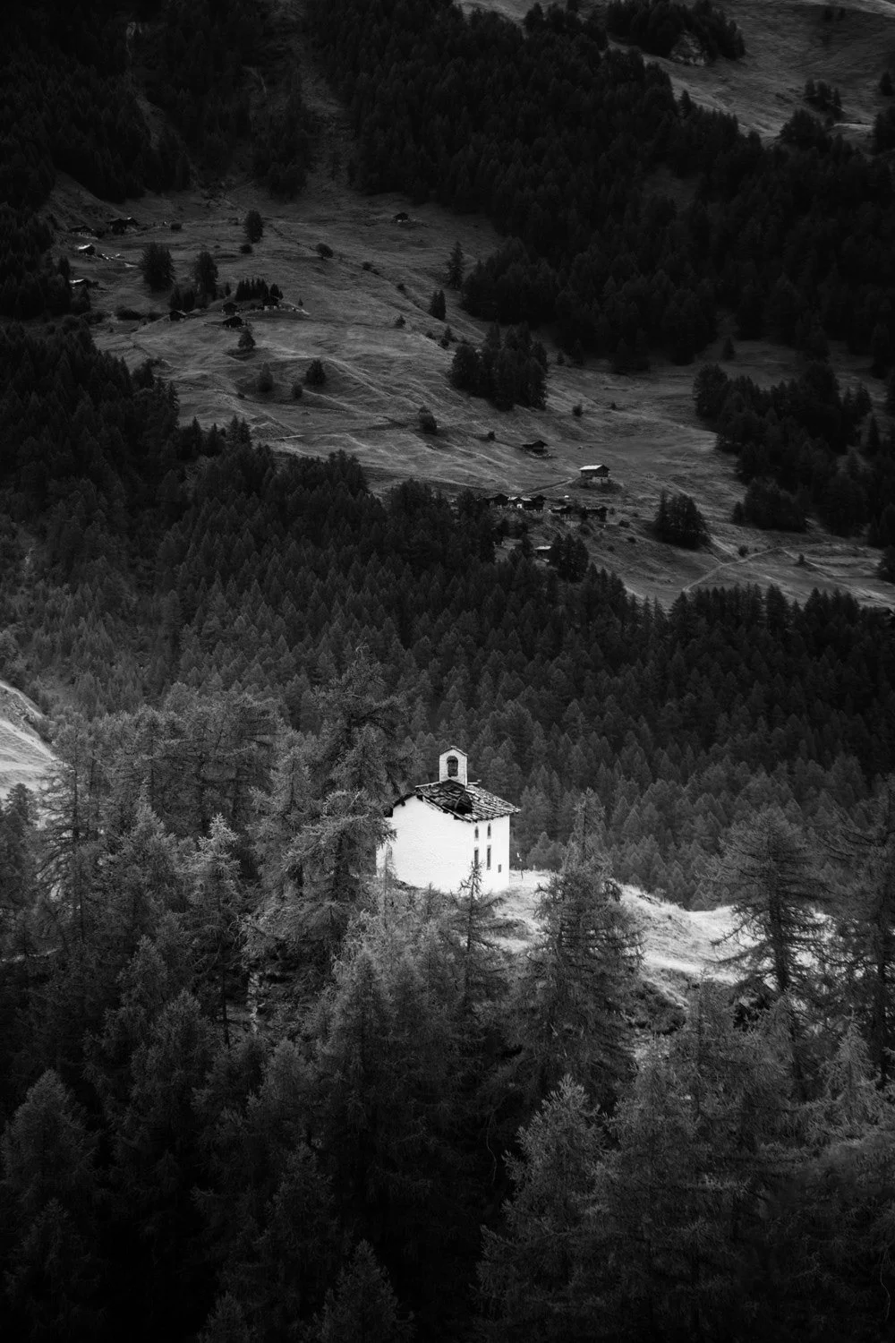 Black and white photograph of a small white church on a hillside surrounded by trees, with a mountainous landscape and scattered houses in the distance. By Auliseo (Aurèle Eliseo Stauffer).