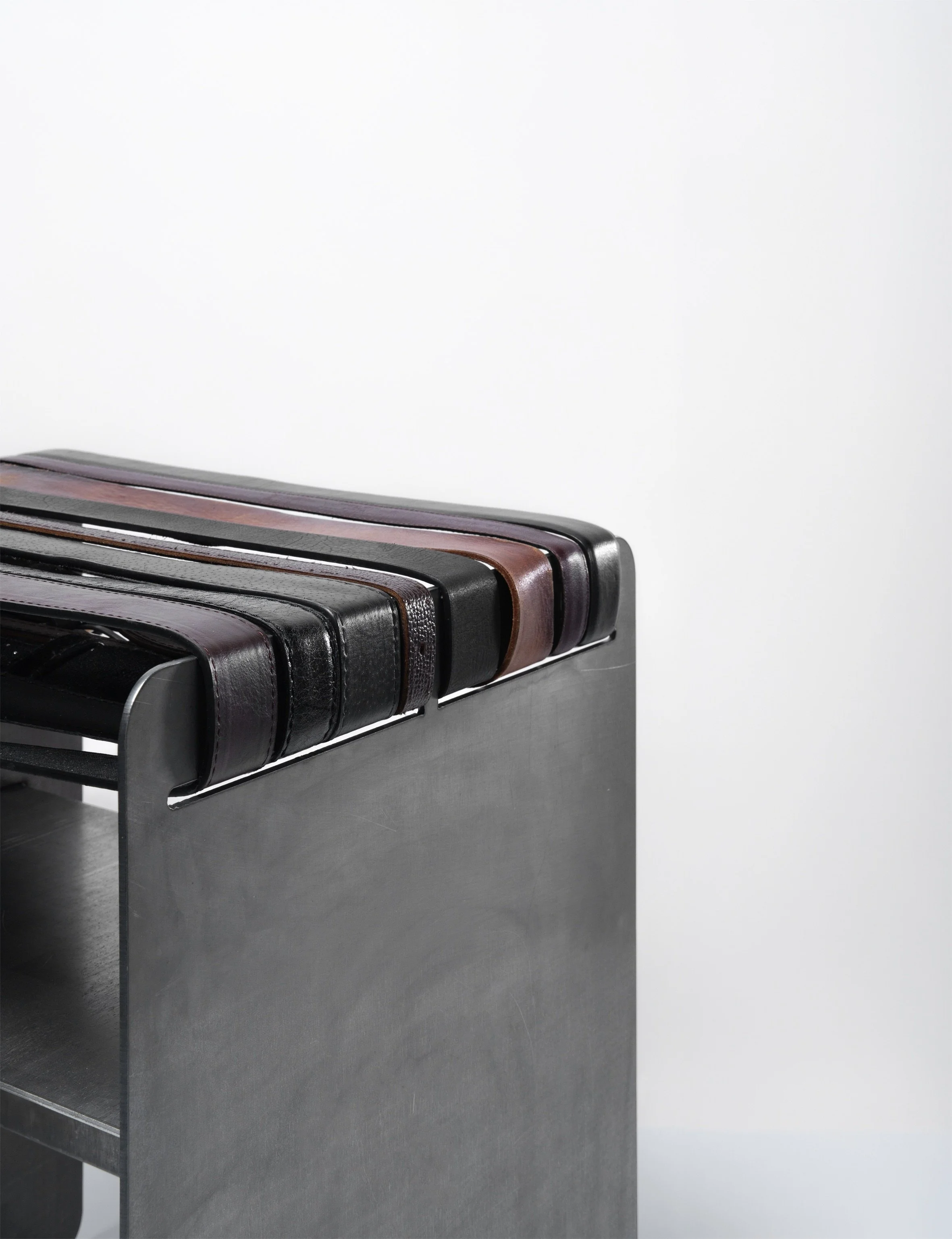 Stacked black, brown, and burgundy leather belts on a steel metal stool against a plain white background. Architecture and Design by Auliseo (Aurèle Eliseo Stauffer).