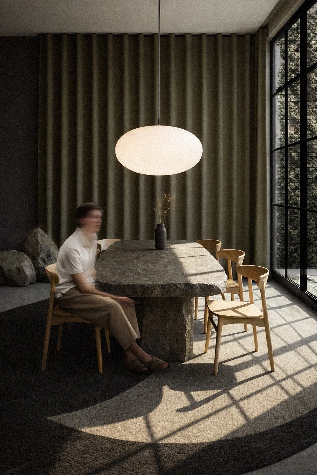 A man sitting on a chair at a large, irregularly shaped stone dining table in a modern room with large windows, vertical curtains, and a hanging pendant light. Interior Design for a Loft called BASE in Zürich by Auliseo (Aurèle Eliseo Stauffer).