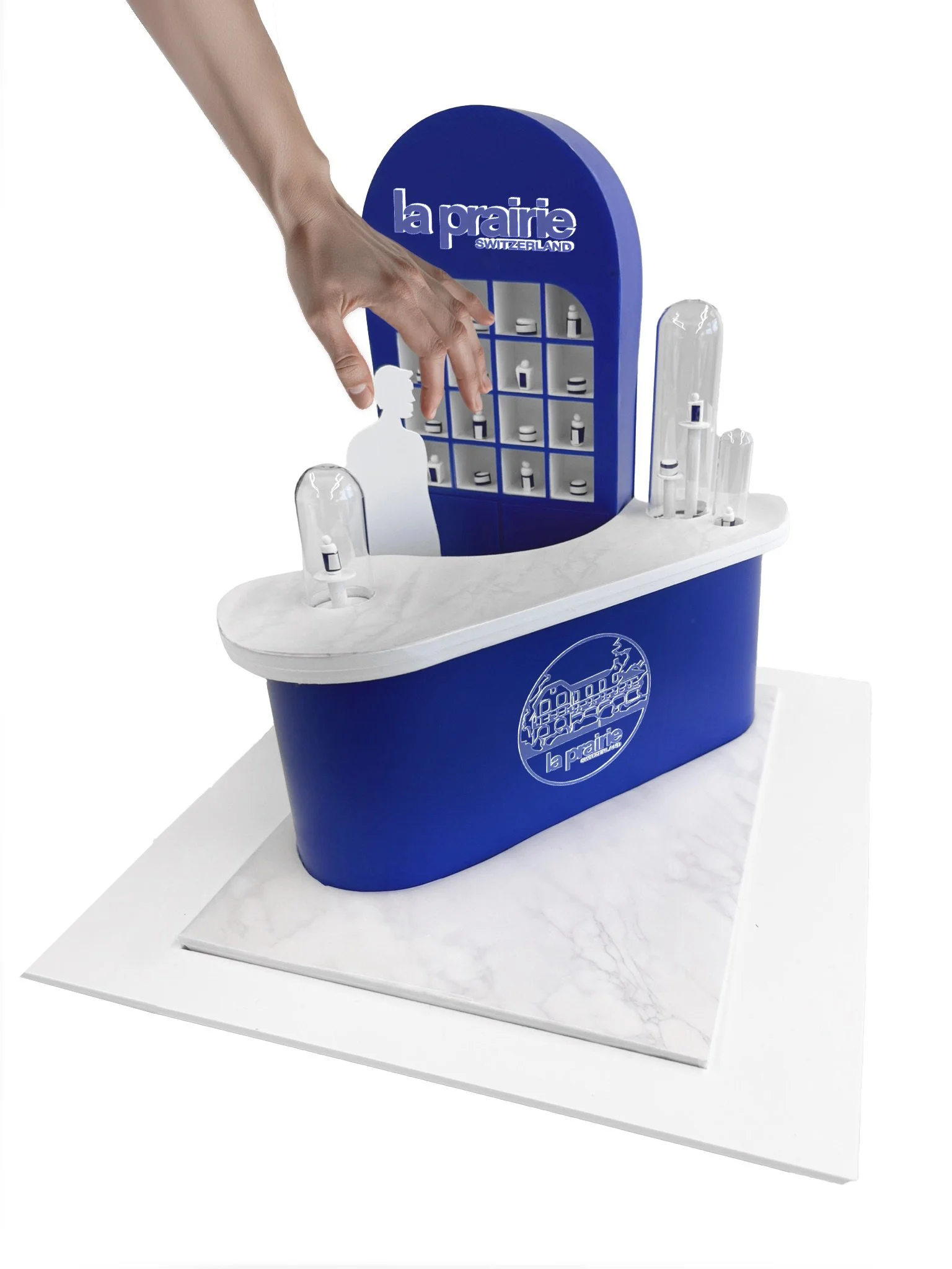 A decorative display with a blue and white La Prairie Switzerland branding, featuring small bottles and containers on a shelf, with a person's hand reaching towards the display.