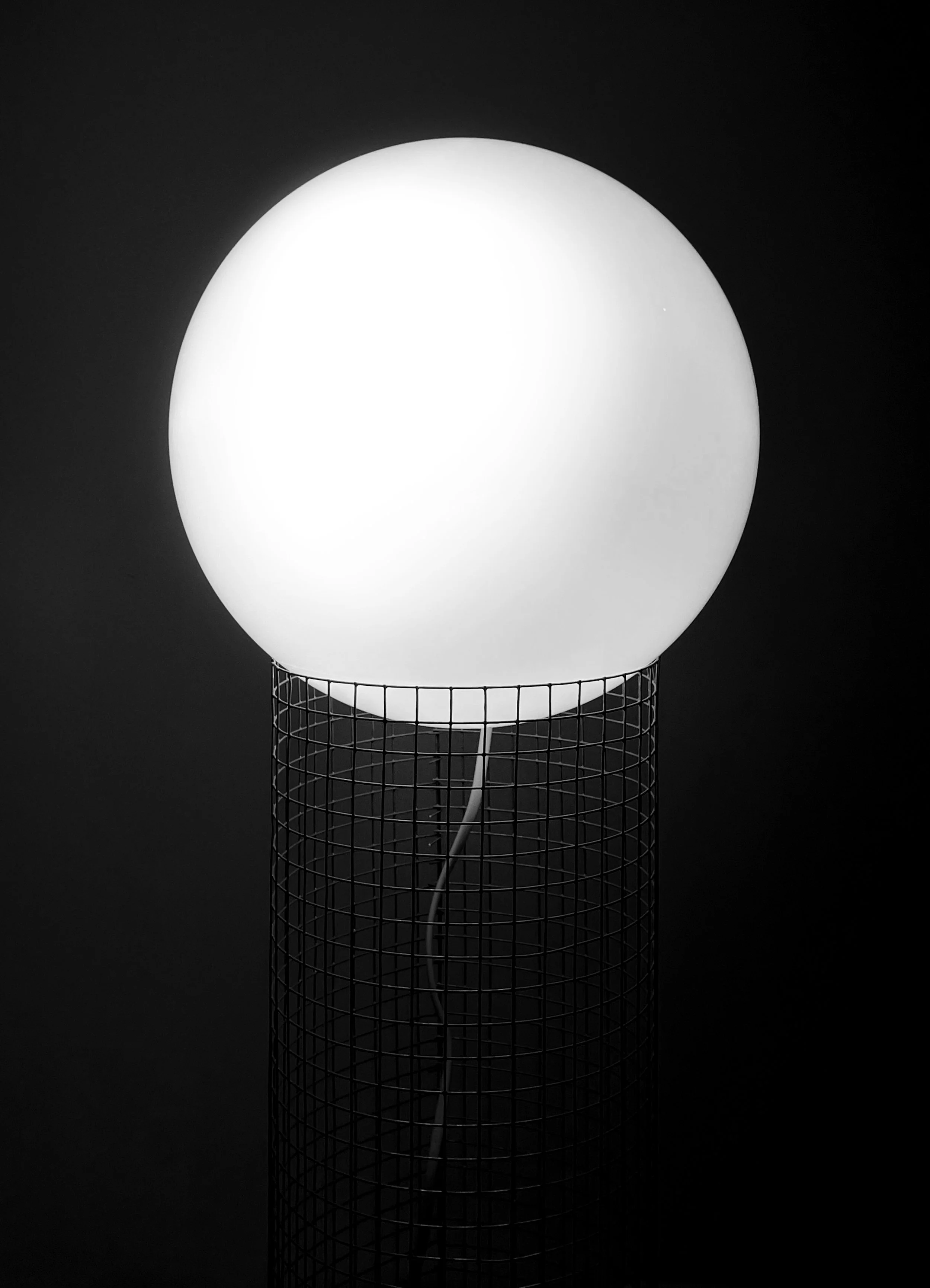 A spherical white lamp in a wire cage lamp shade against a dark background. Architecture and Design by Auliseo (Aurèle Eliseo Stauffer).