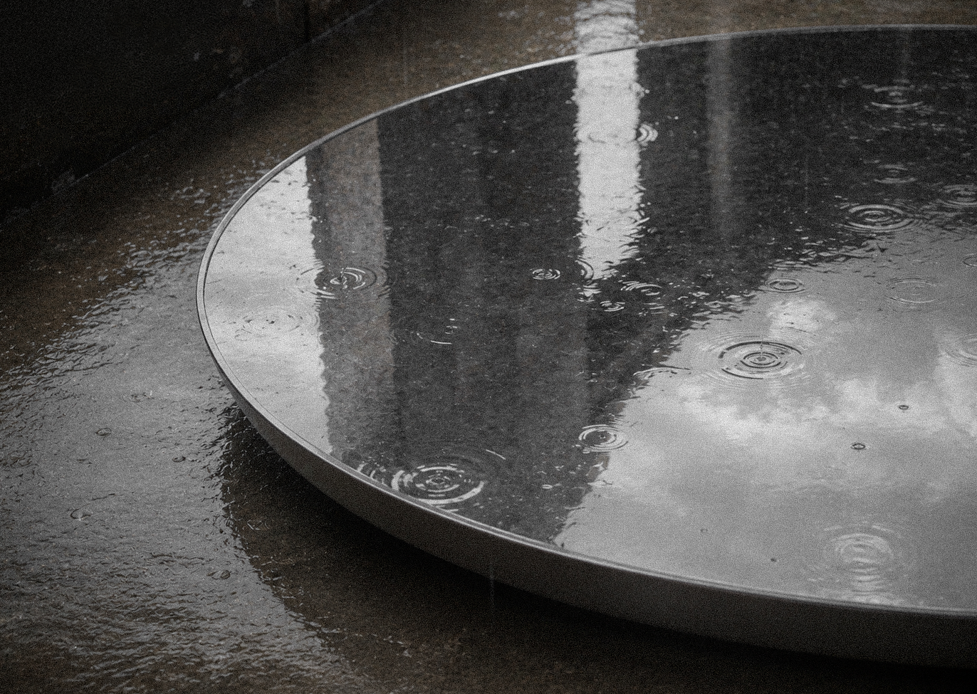 A close-up of a round full basin on a wet, dark surface, reflecting sky and through rain a Chapel. By Auliseo (Aurèle Eliseo Stauffer).