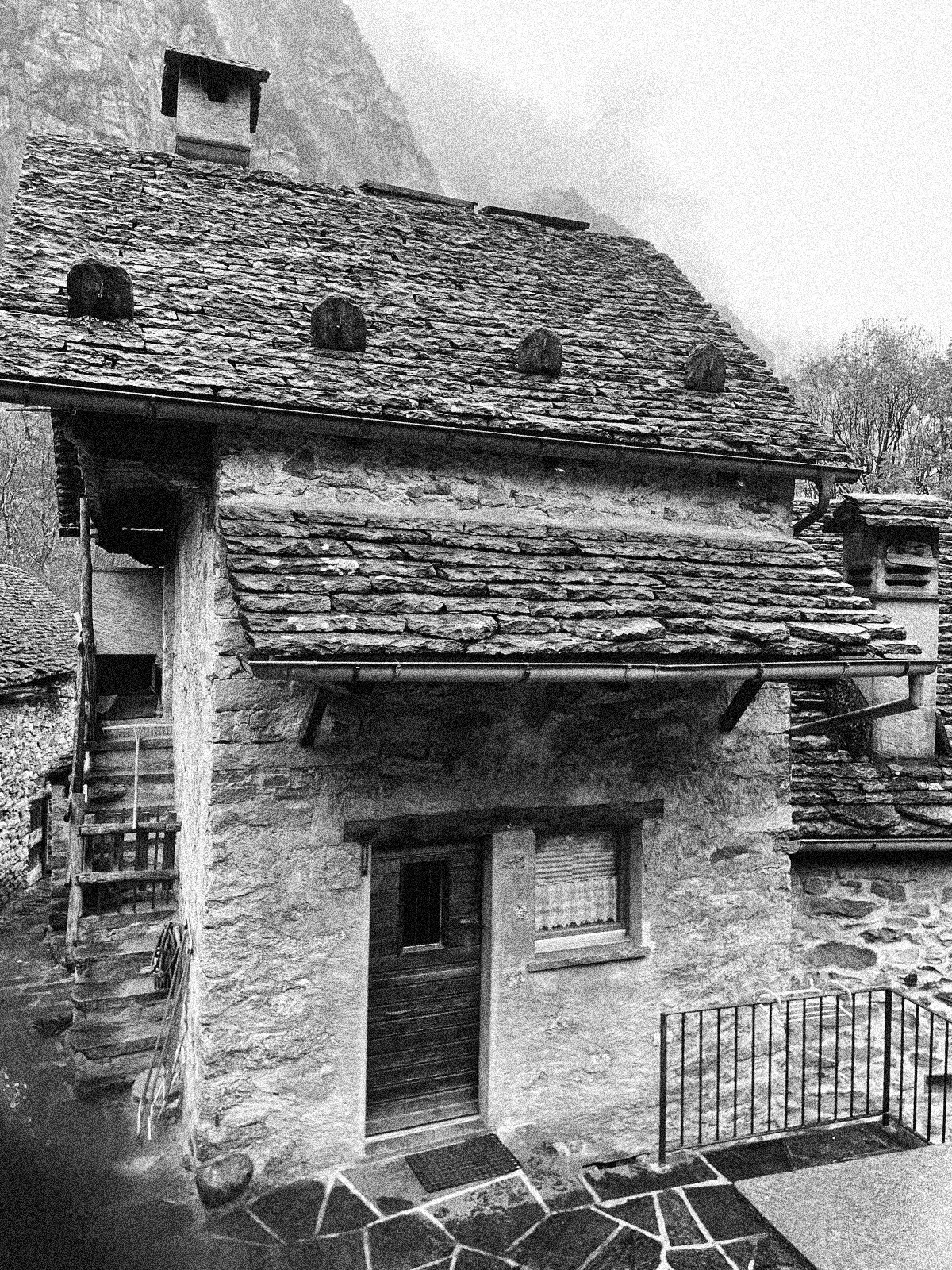 Black and white photo of a rustic stone house with a sloped slate roof, small windows, and a wooden door, surrounded by a stone-paved courtyard and a metal railing.