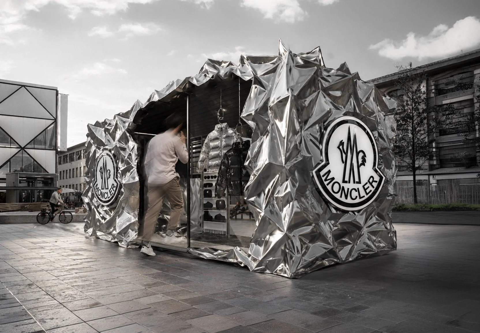 A Moncler pop-up store in a city square with a reflective, metallic exterior and large Moncler logos on the sides. A person is looking inside, and a cyclist is passing by. Architecture and Design by Auliseo (Aurèle Eliseo Stauffer).