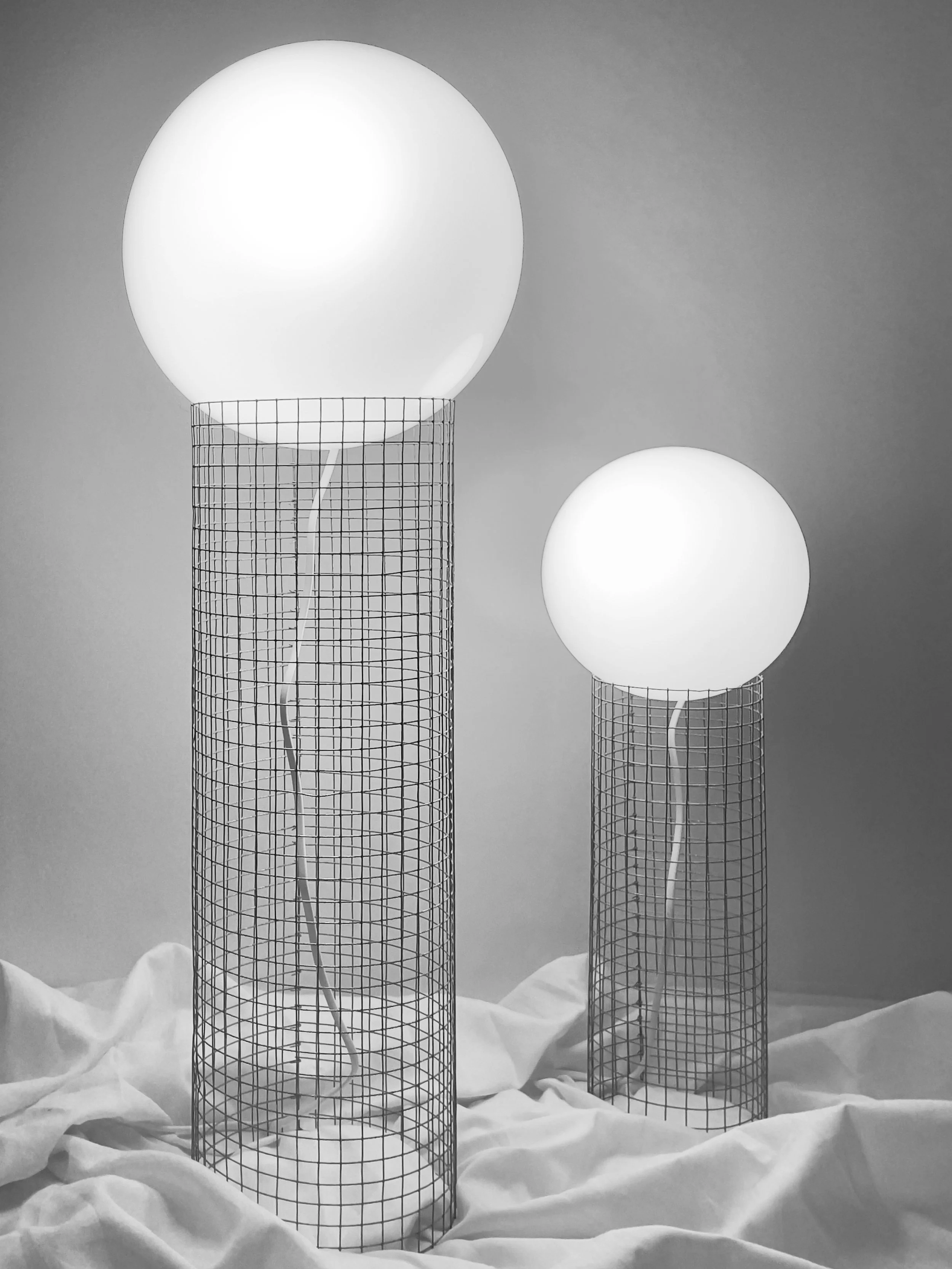 Black and white photo of three tall wireframe cylindrical lamps with round bright white glass bulbs on top, arranged in a staggered line against a plain background, with crumpled white fabric at the bottom. Architecture and Design by Auliseo (Aurèle
