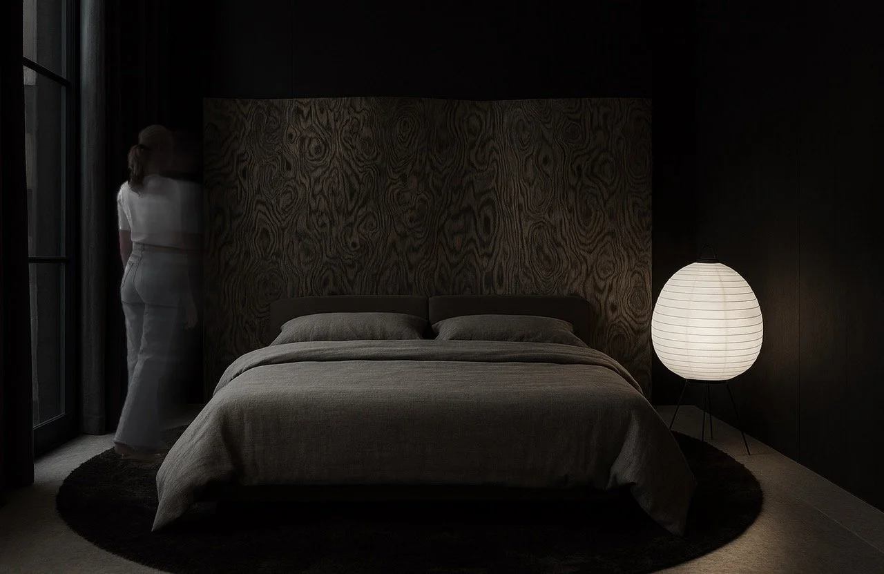 A dimly lit bedroom with a neatly made bed, a large white paper lantern lamp, against a dark wall with a textured headboard, and a woman in white standing near a window with curtains.  Interior Design for a Loft called BASE in Zürich by Auliseo.