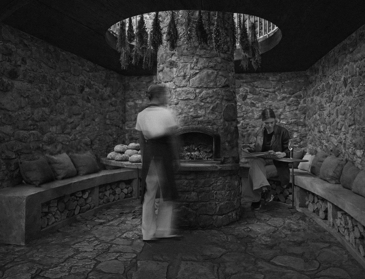 Black and white photo of a cozy stone-walled room with a central stone fireplace, bench seating with cushions on three sides, a person sitting at a small table drinking wine and reading, and a server standing near the fireplace, which has hanging herbs or plants from the ceiling opening.