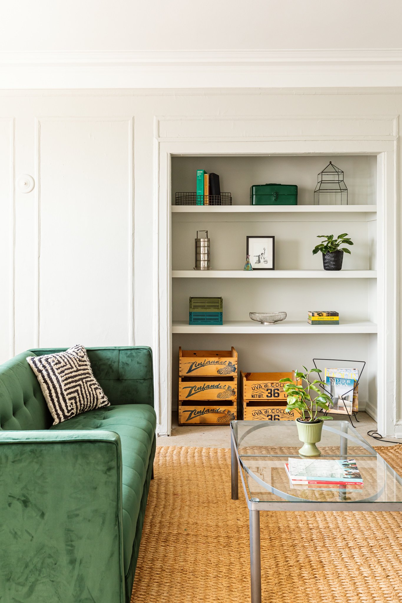 Living room with a green sofa, a glass coffee table with a potted plant, and built-in white shelves with books and decorative items.
