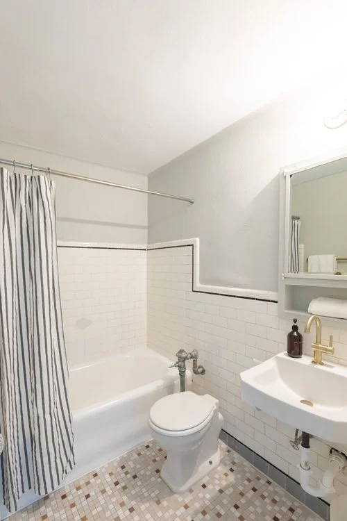 Small bathroom with white tile walls, a bathtub with a shower curtain, a toilet, and a small white sink with a mirror and shelf above.