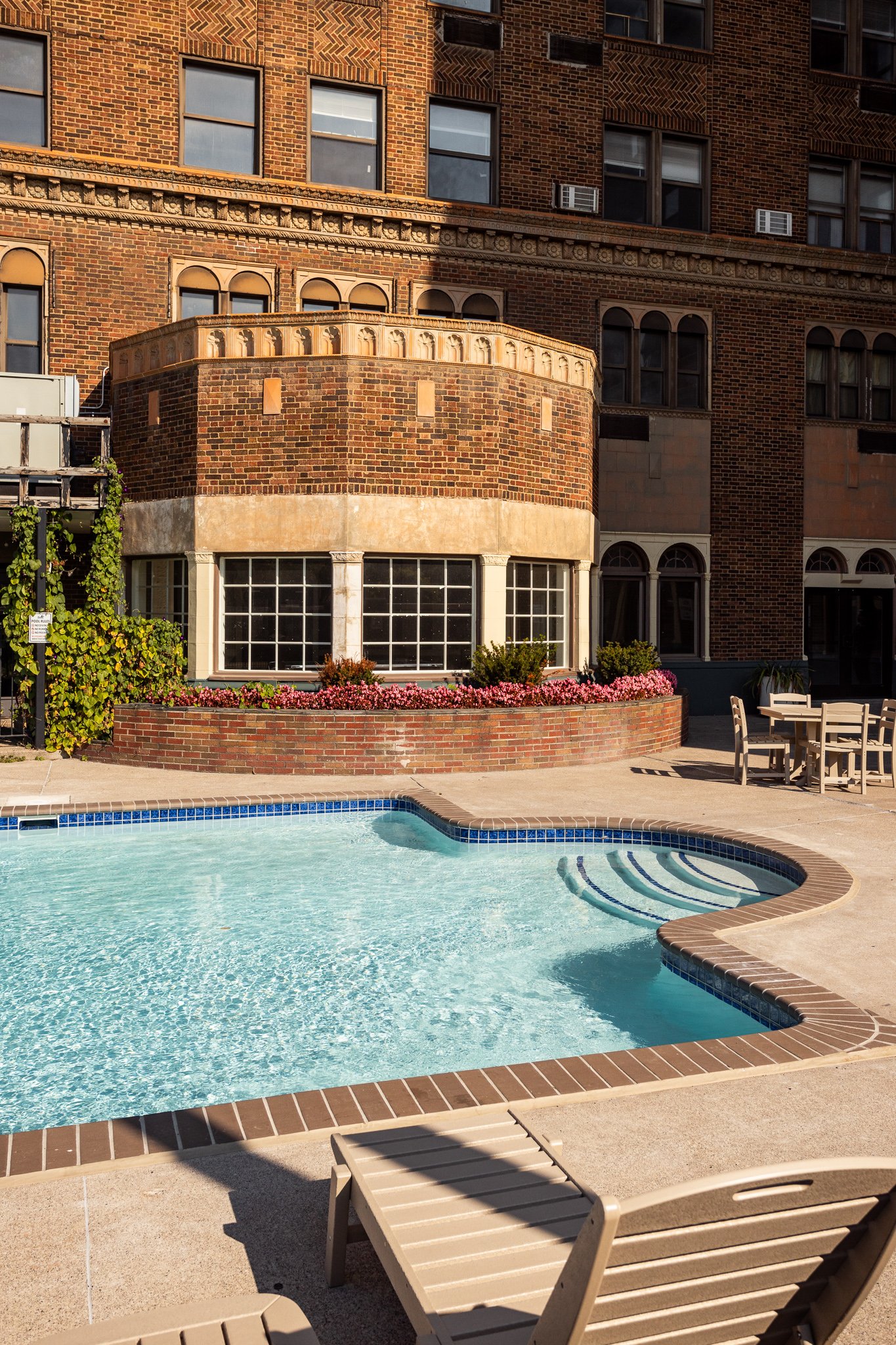 A swimming pool with clear water, surrounded by a concrete deck, with a brick building and outdoor furniture in the background.