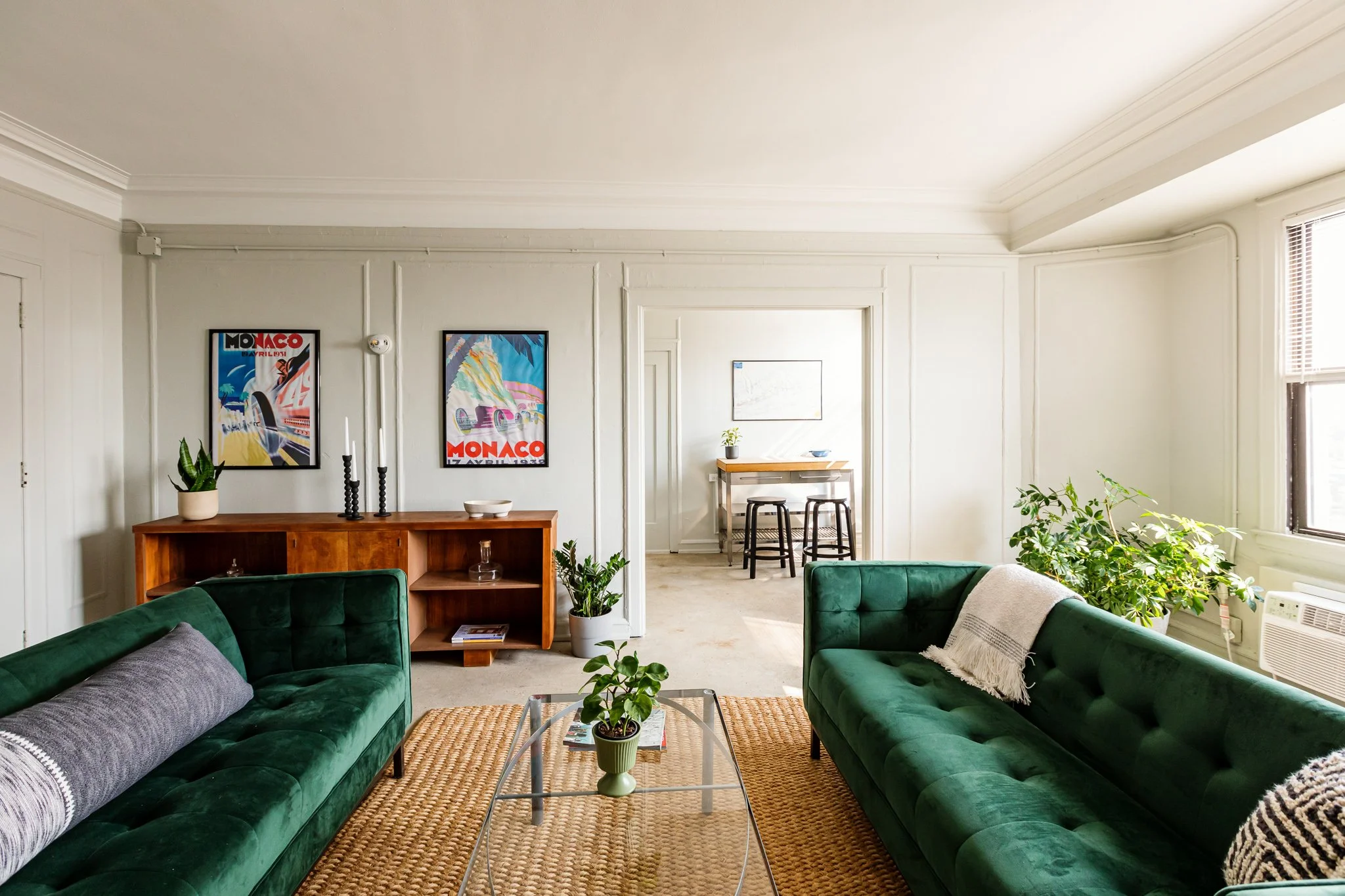 Living room with two green velvet sofas, a glass coffee table with a small potted plant, and a wooden credenza with decorative items. Artwork with vintage racing posters hangs on the wall. Near the window, there are large plants. In the background, t