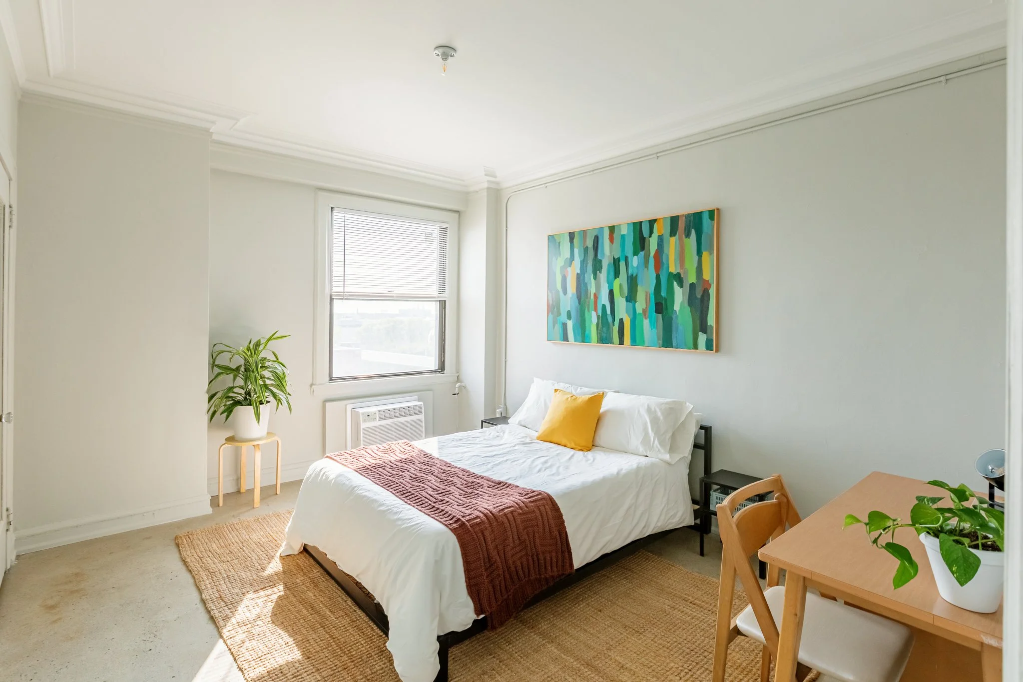A bright bedroom with a bed, a window with blinds, a plant on a table, a colorful abstract painting on the wall, a small desk with a plant, and a beige rug.