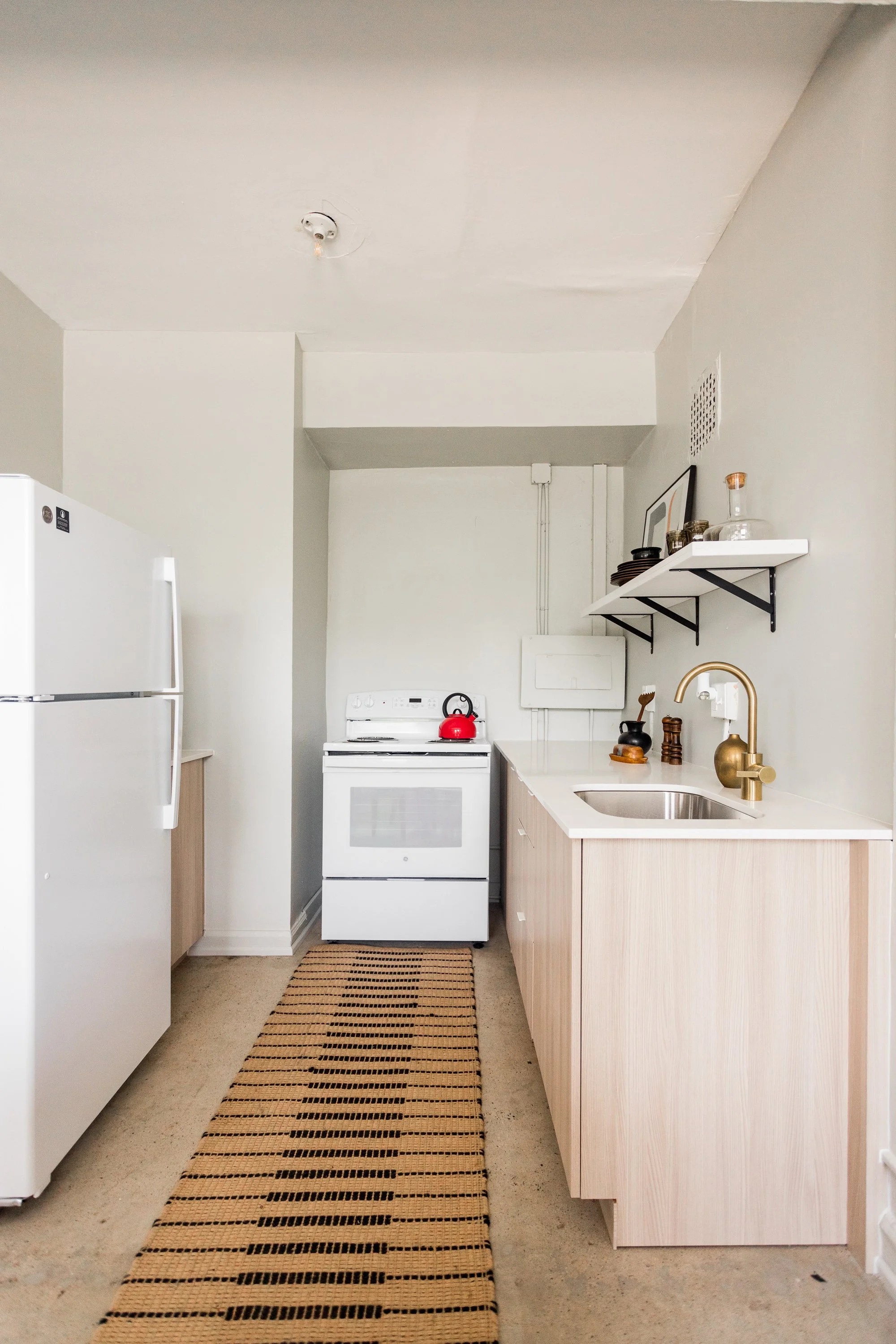 Minimalist kitchen with white refrigerator, white stove with red kettle, light wood cabinets, white countertop, black wall shelf with decorative items, gold faucet, and beige woven rug on concrete floor.