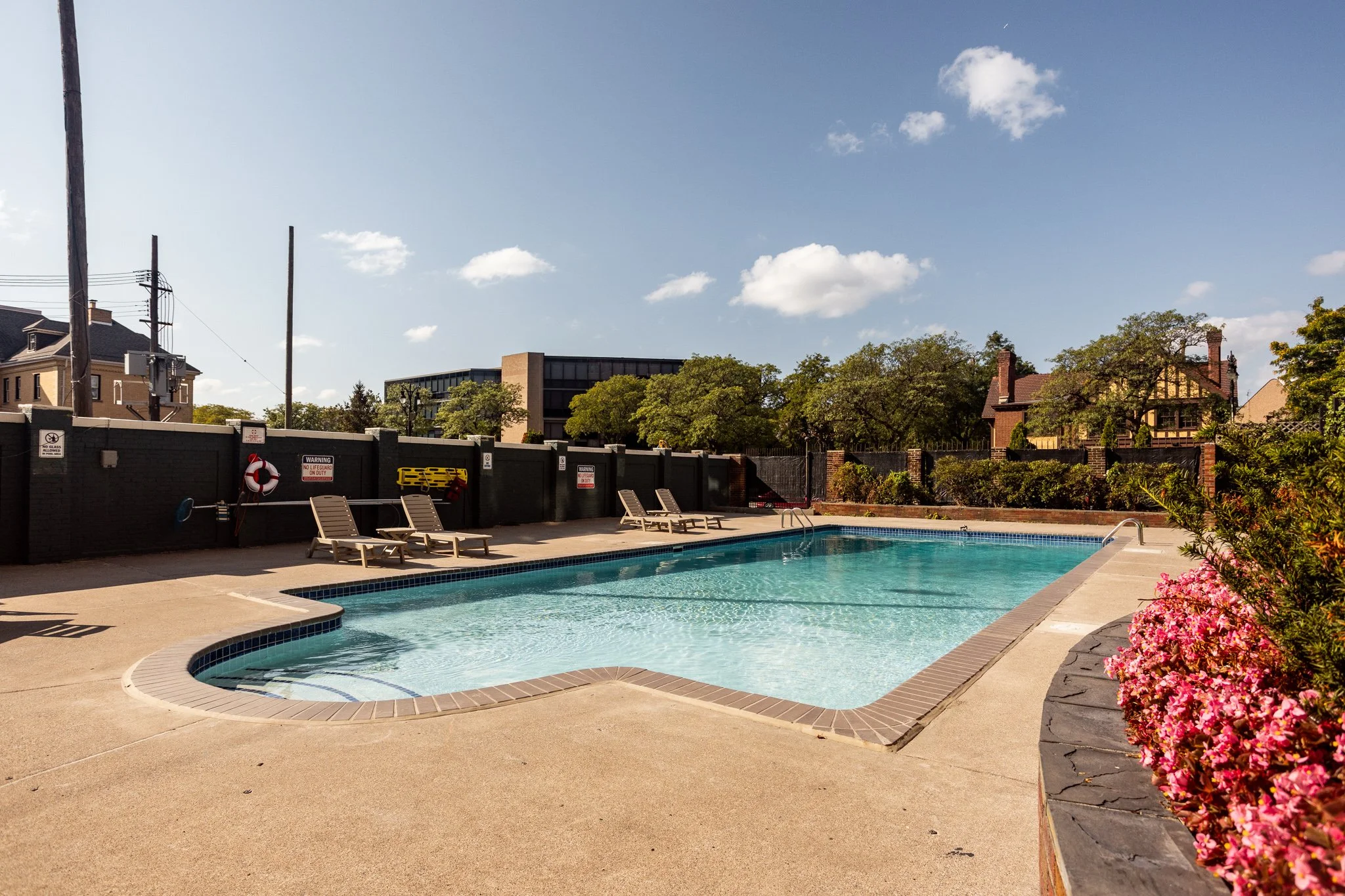Outdoor swimming pool with lounge chairs beside it, surrounded by a brick border and landscaping, under a partly cloudy sky.