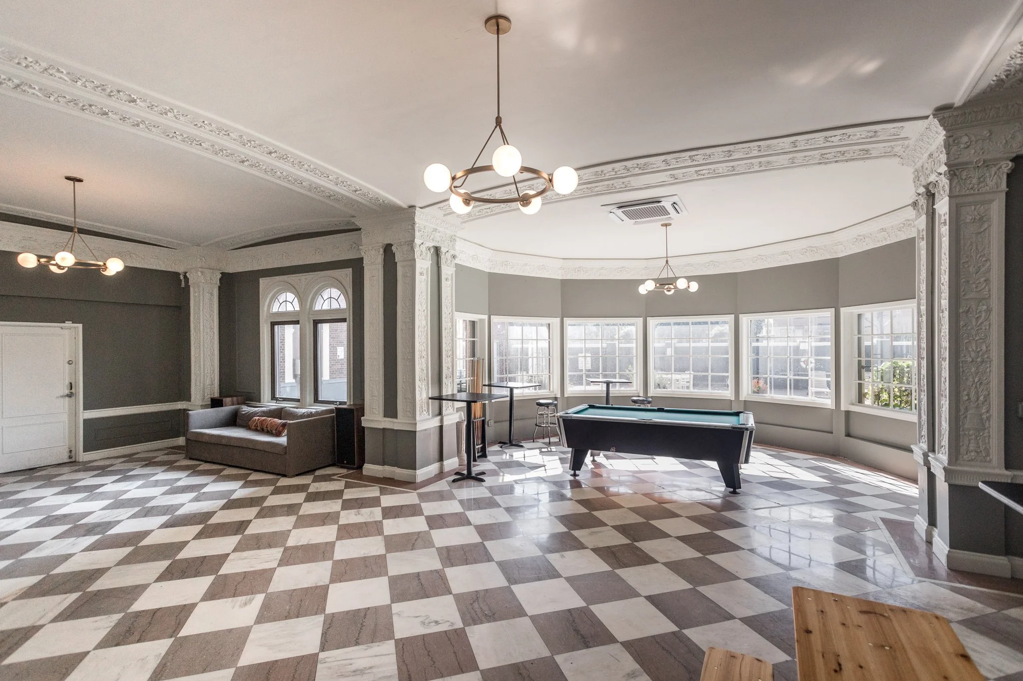 Spacious room with checkered floor, gray walls, decorative white crown molding, large bay windows, a pool table, a gray sofa, and modern ceiling lighting fixtures.