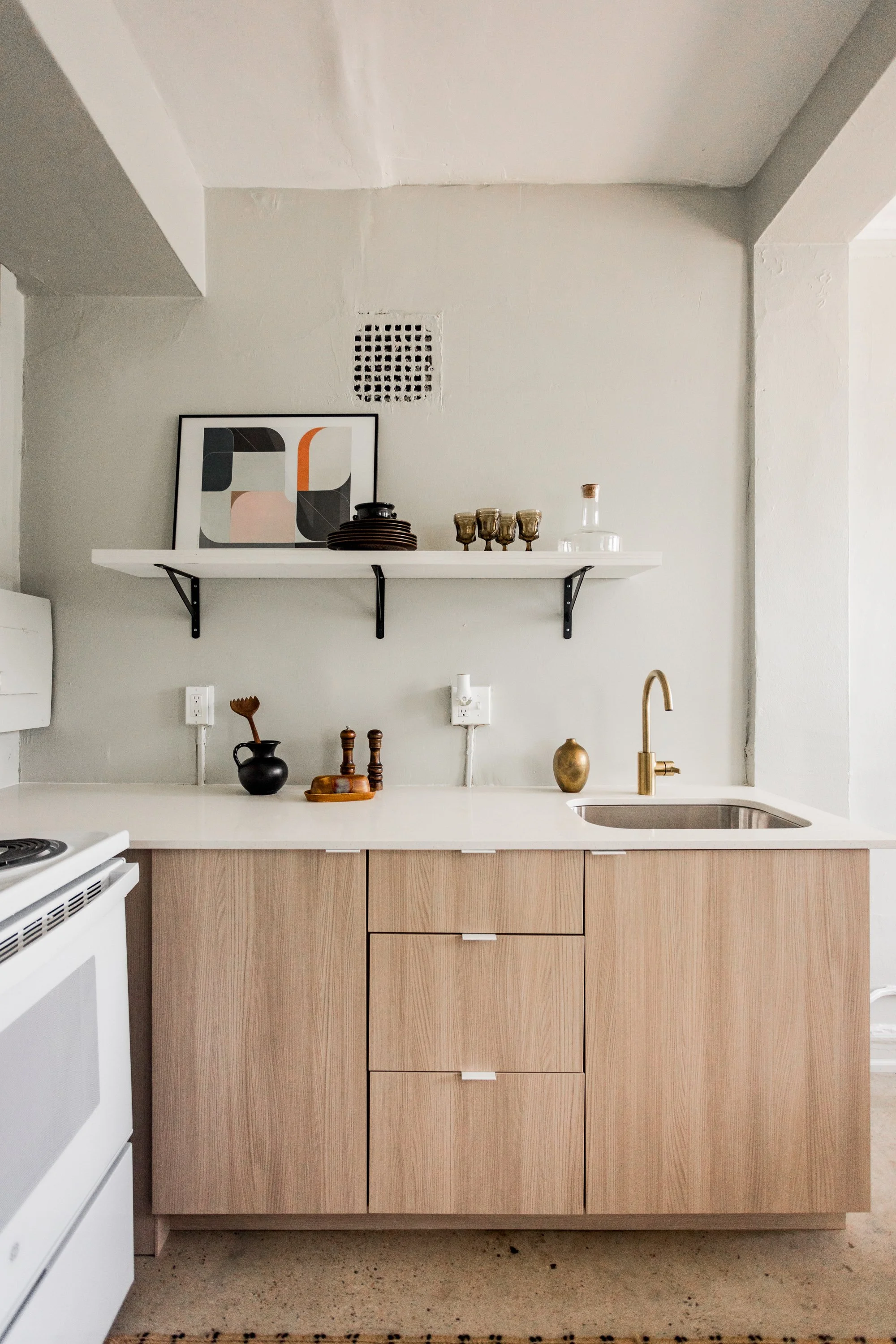 Minimalist kitchen with a light wood cabinet, white countertop, brass faucet, open shelf with decorative items, and abstract artwork on the wall.