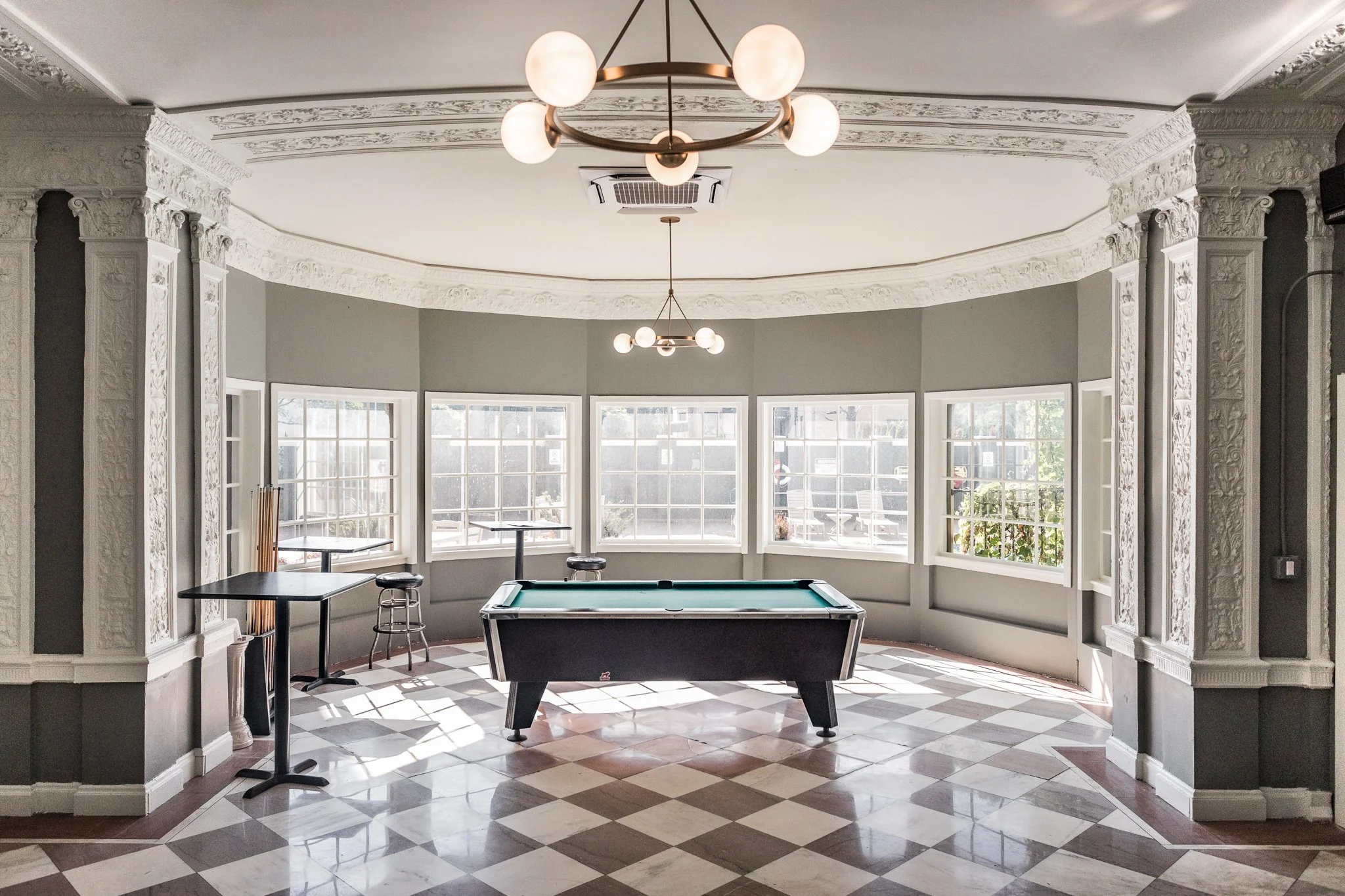 An interior room with a gray color scheme, ornate white crown molding, and a black pool table in the center. The room has large windows surrounding it, with sunlight streaming in, and features a modern chandelier with globes hanging from the ceiling.