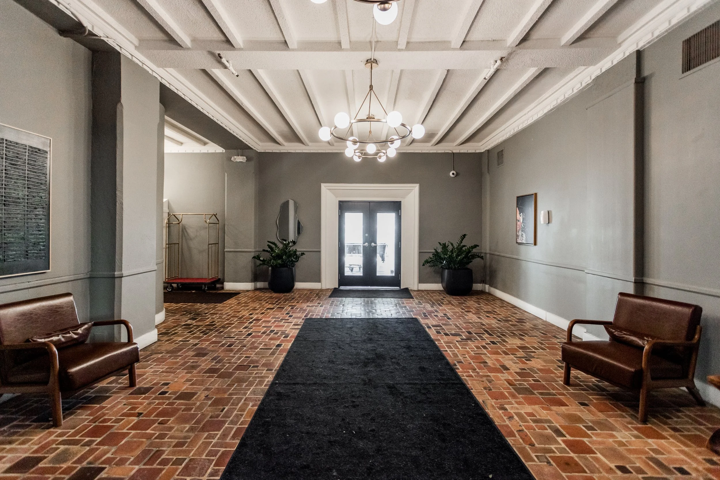 Empty lobby with brick floor, gray walls, two brown leather benches, black rug, black double doors, two potted plants, hanging chandelier, and artwork on walls.
