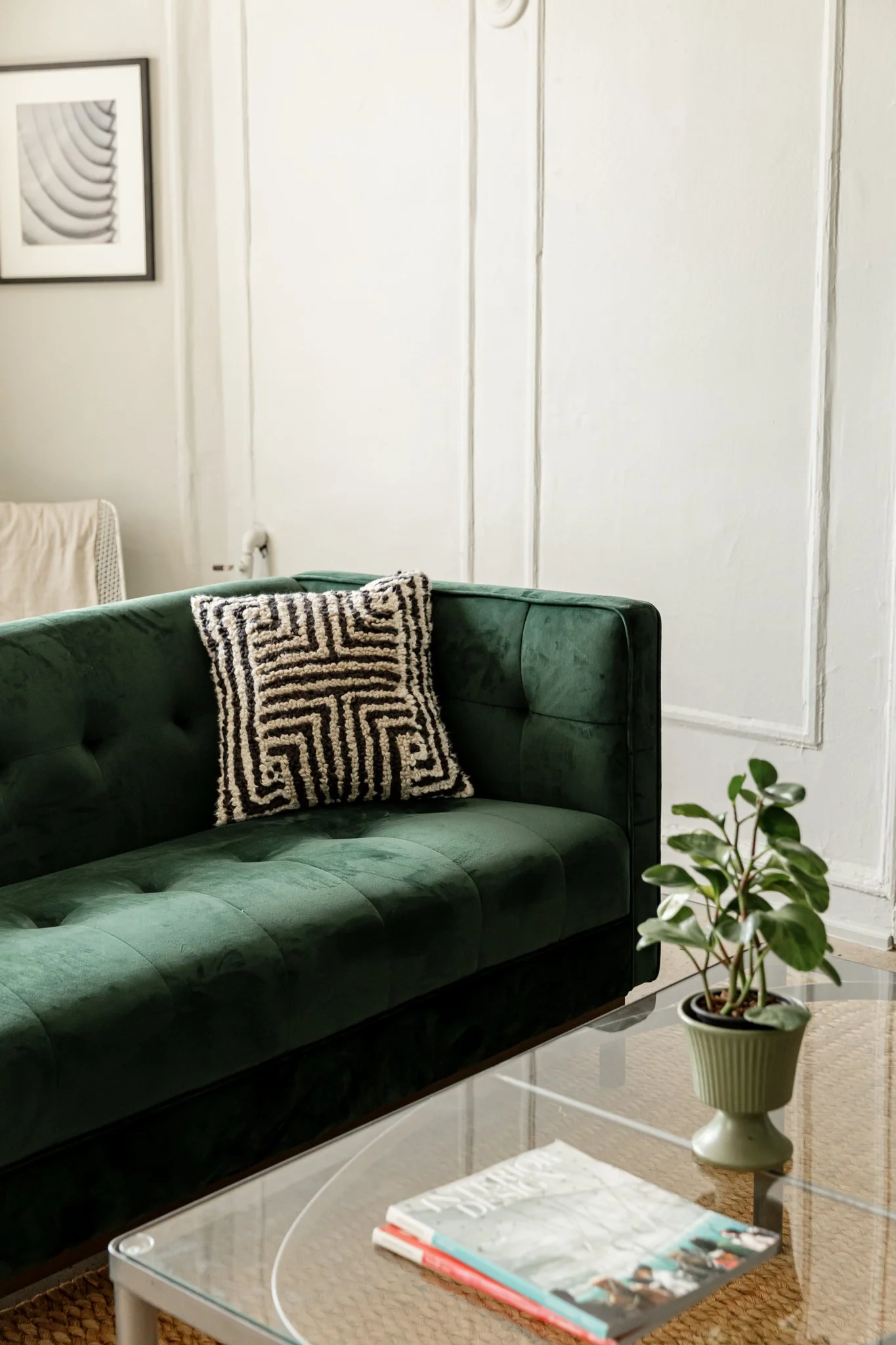 A living room with a dark green velvet sofa, decorative pillow, a potted plant, a glass coffee table with magazines, and framed artwork on the wall.