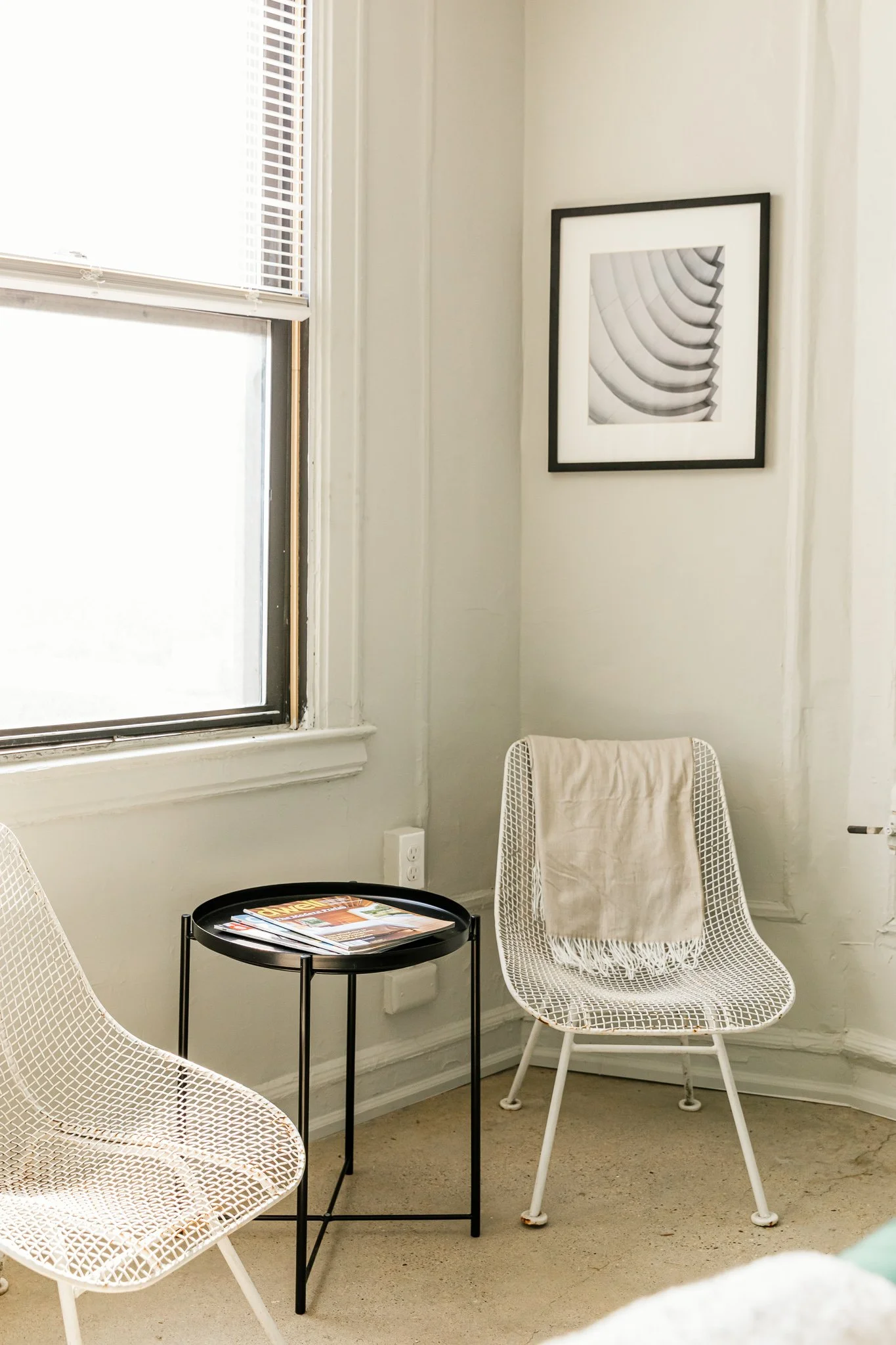A cozy corner with two white wire chairs, a black round side table with magazines, a window with blinds, and a black-and-white framed art print on the wall.