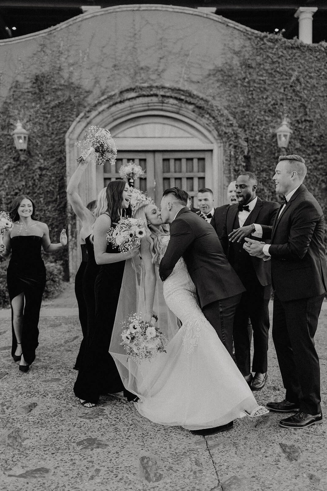 Black and white photo of groom kissing bride with wedding party in the background 