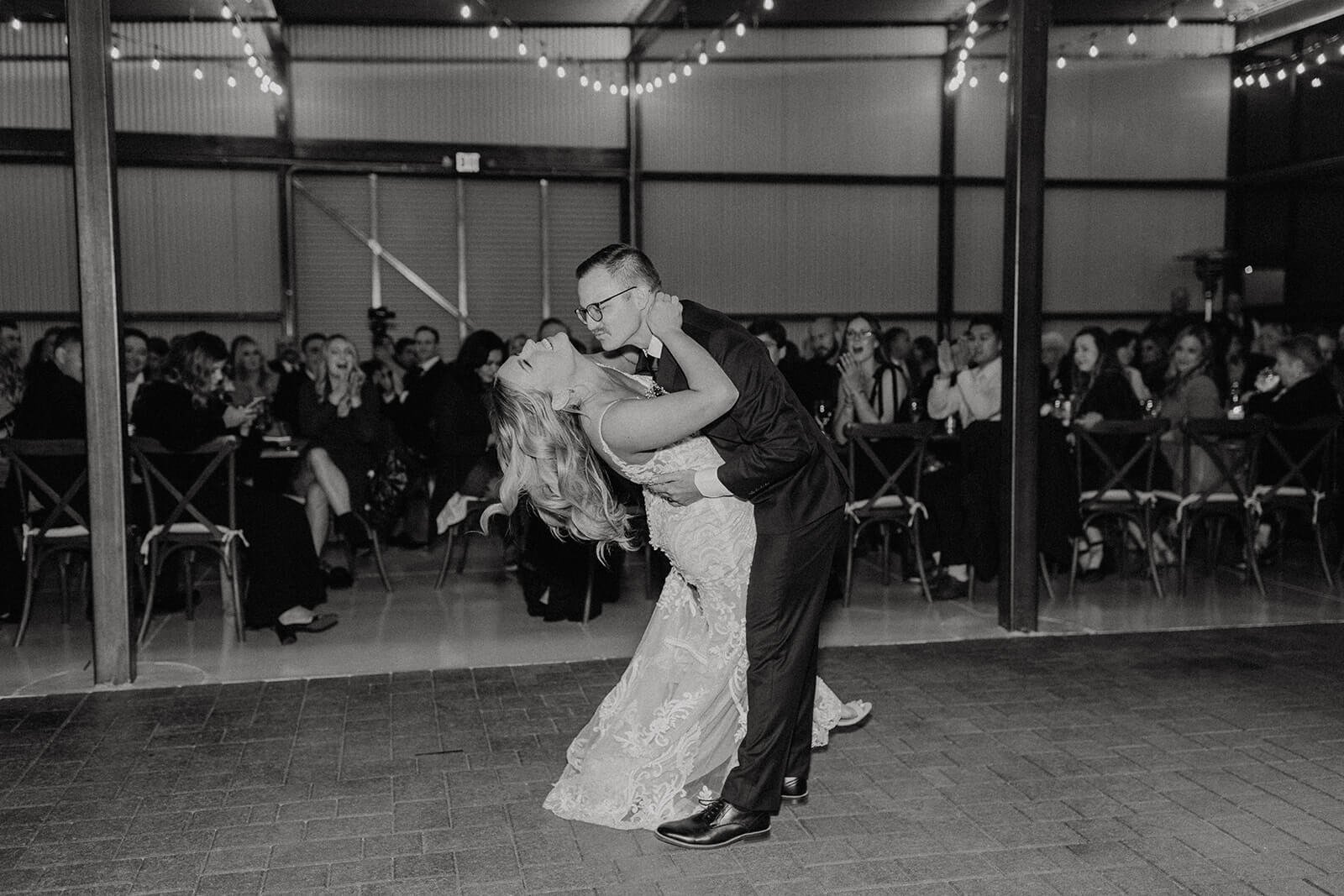 Black and white photo of groom dipping bride during dance with market lights strung above