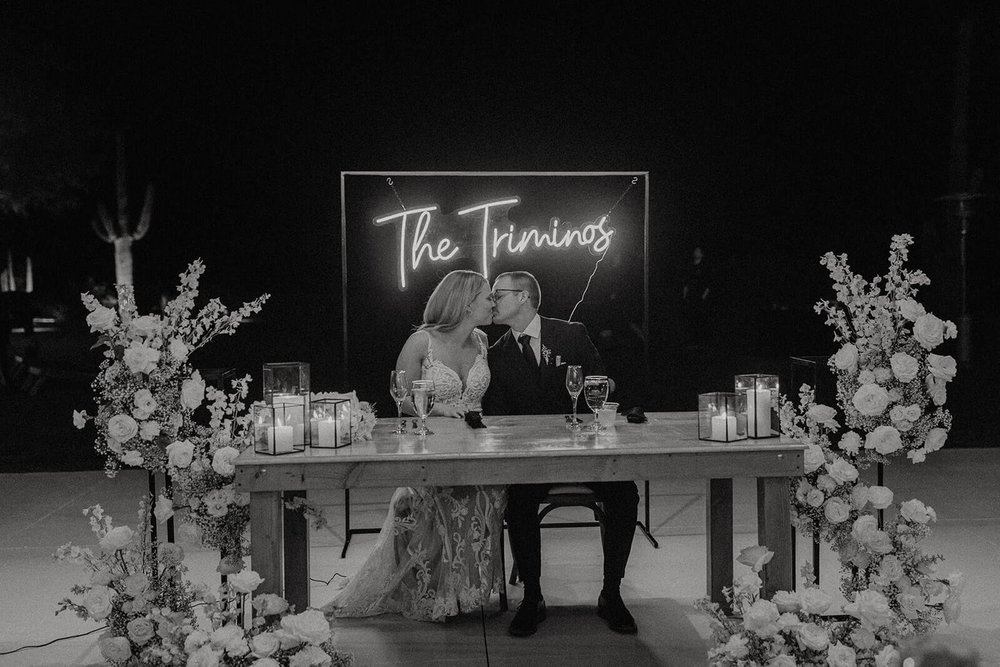 Bride and groom kiss at head table with fluorescent scripted light of their last name behind them 