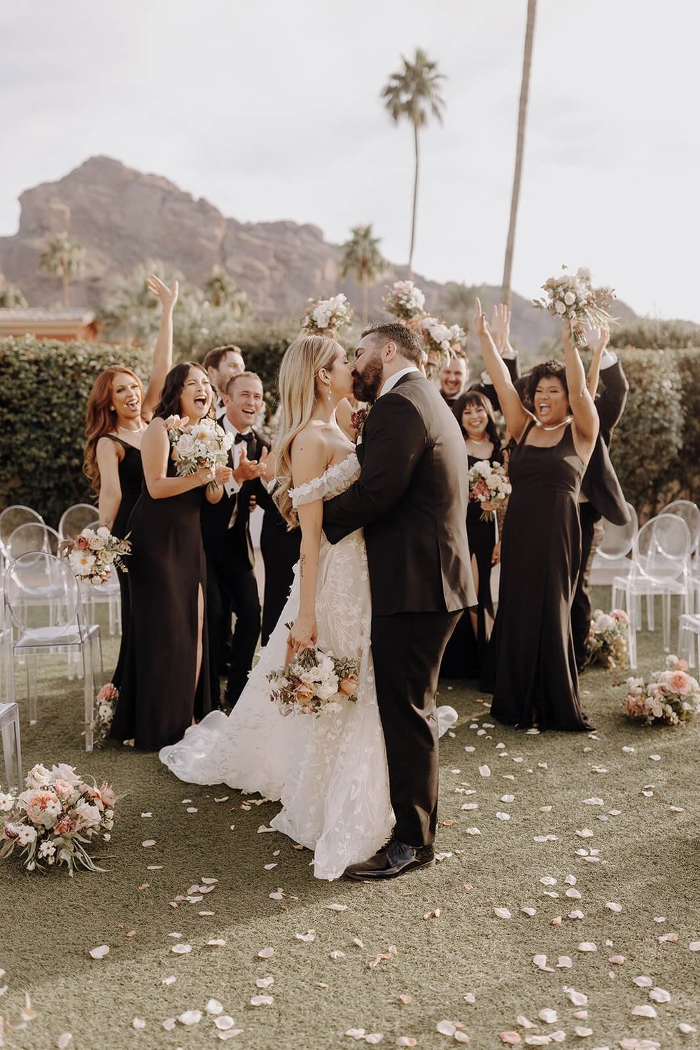 Bride and groom kissing outside with bridal party around them, palm trees and mountains in the background