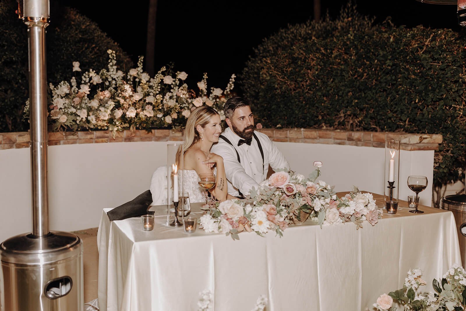 Bride and groom listening to toasts with floral arrangements behind them at outdoor luxury wedding reception