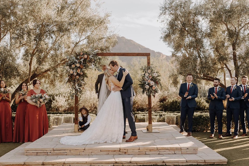 Bride and groom first kiss under arbor in outdoor desert wedding, mountains in the background