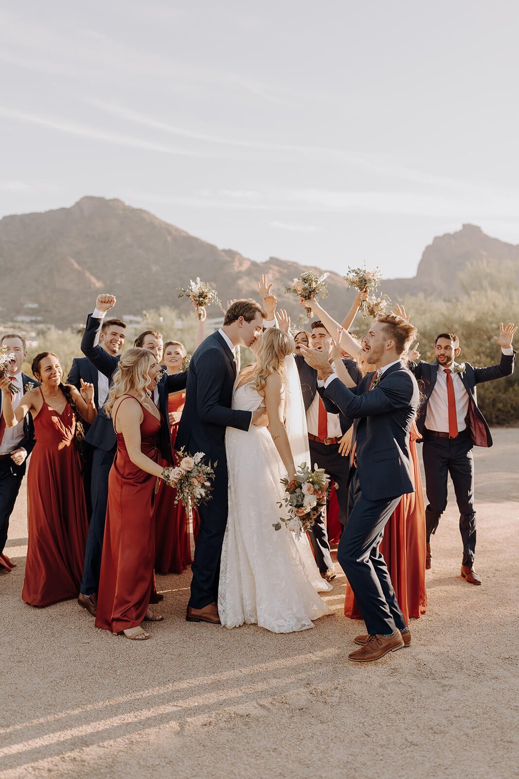 Bride and groom kissing with bridesmaids and groomsmen celebrating around them, desert mountains in the background