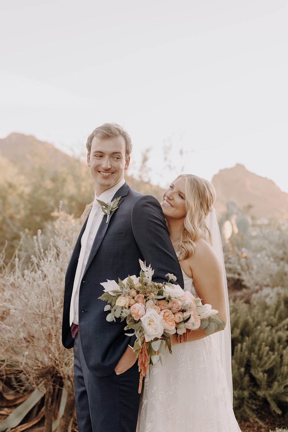 Bride and groom standing outside. Bride standing behind groom looking at him, groom looking back at bride