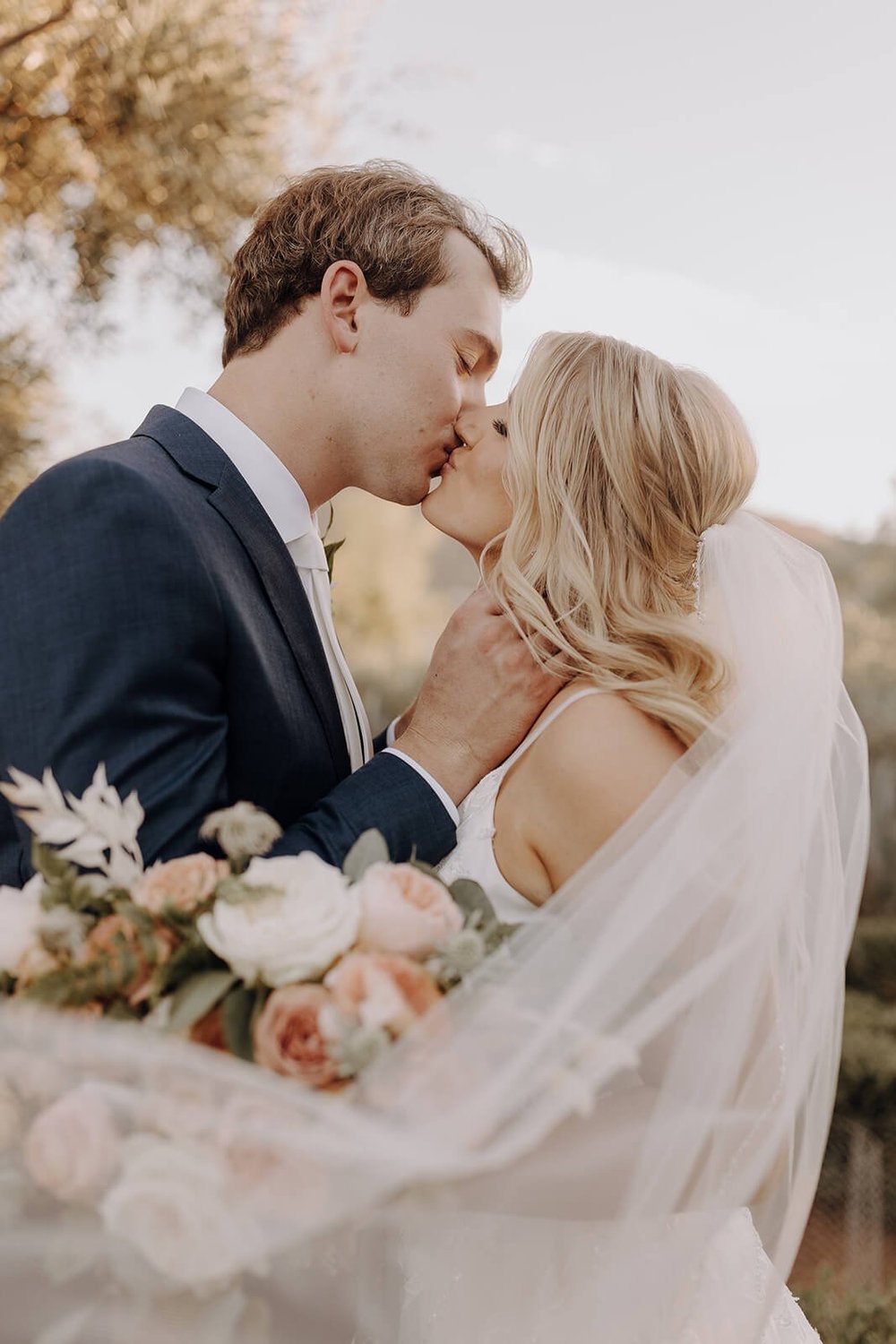 Bride and groom kissing, bride holding bouquet, bride's veil around them