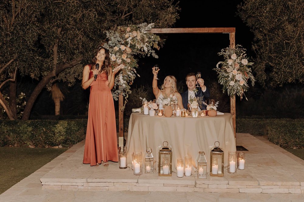 Bride and groom seated under arbor at evening outdoor desert wedding reception, toast to speeches