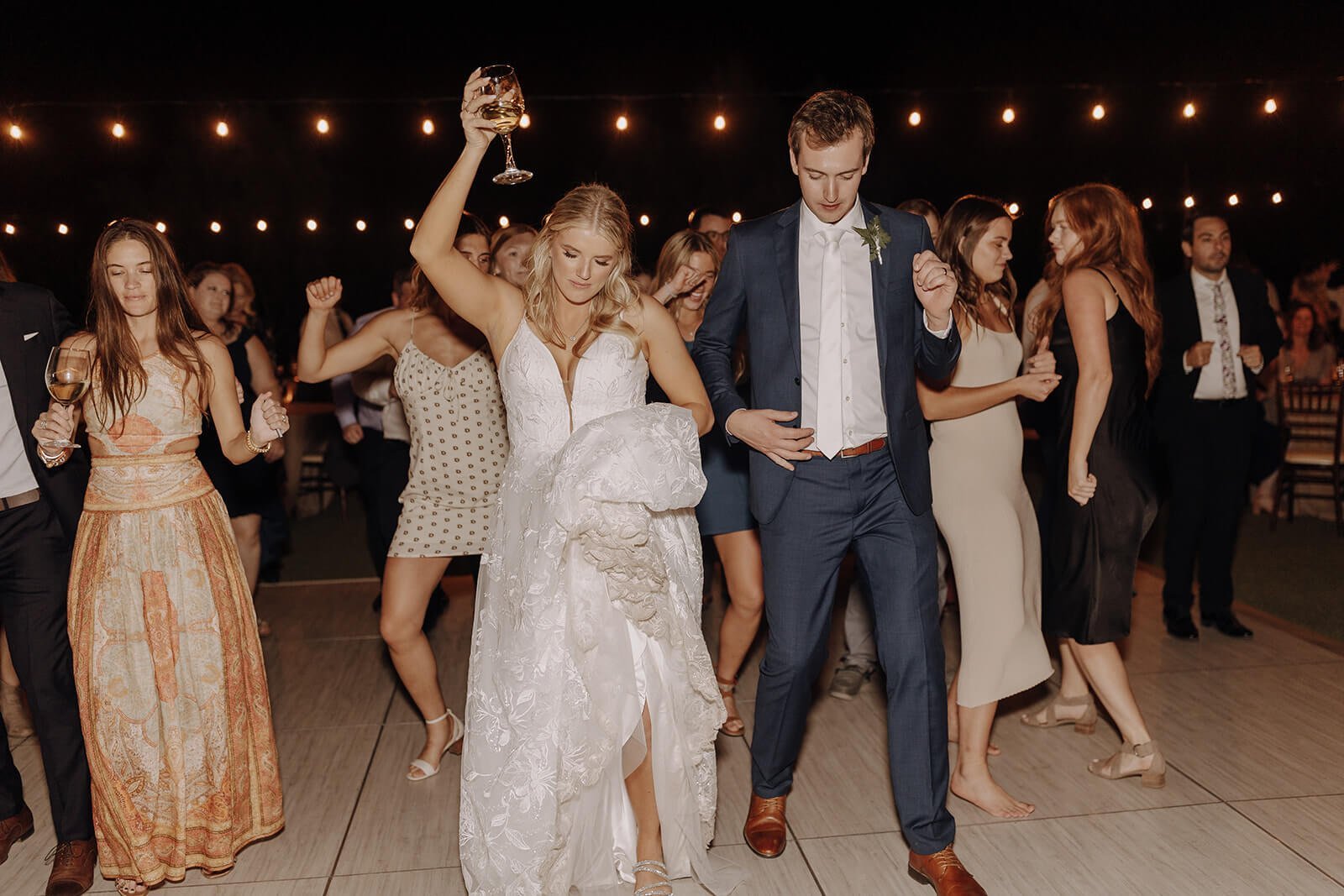 Bride and groom dance with guests under lights at an evening outdoor desert wedding reception
