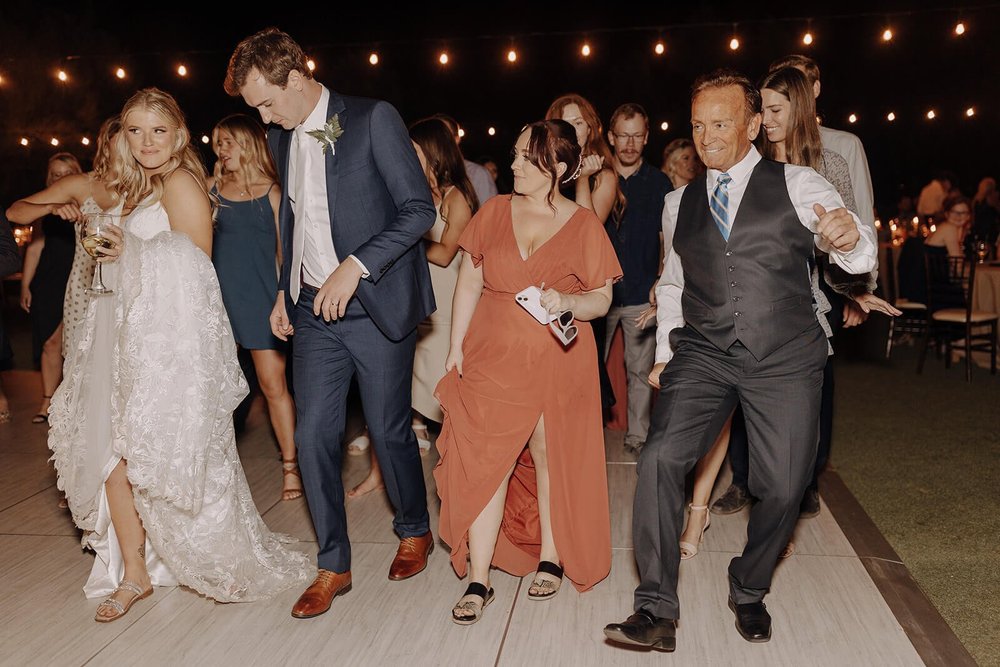 Bride and groom dance with guests under lights at an evening outdoor desert wedding reception