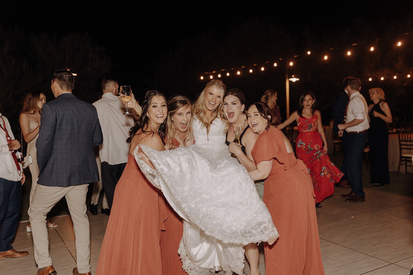 Guests lift bride on dance floor under lights at outdoor evening desert wedding reception