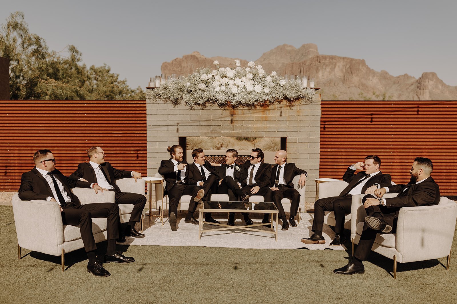 Groom and groomsmen in black suits sitting on outdoor furniture at a desert wedding venue in Arizona