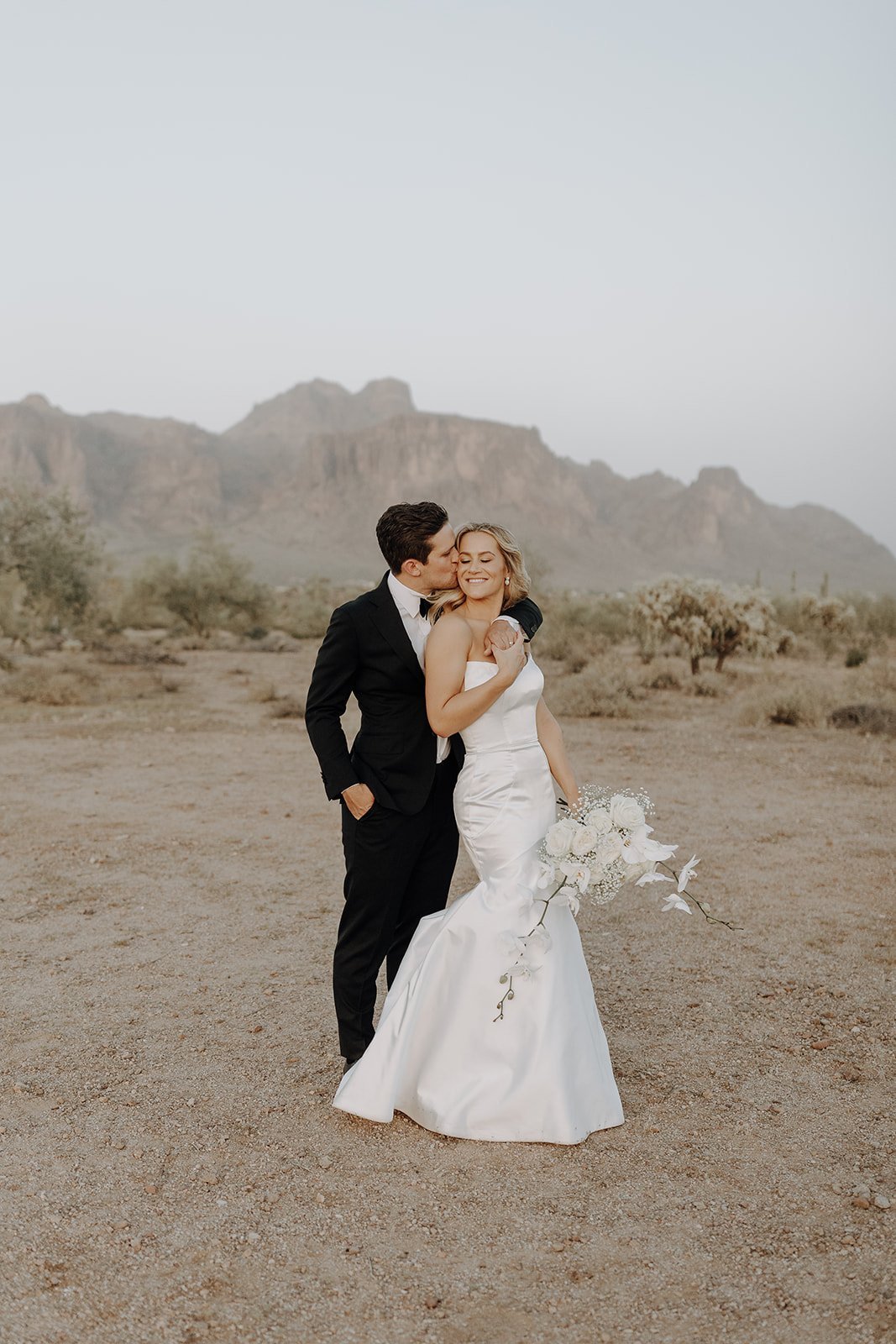 Groom kisses bride on the cheek during couple photos at their desert wedding 