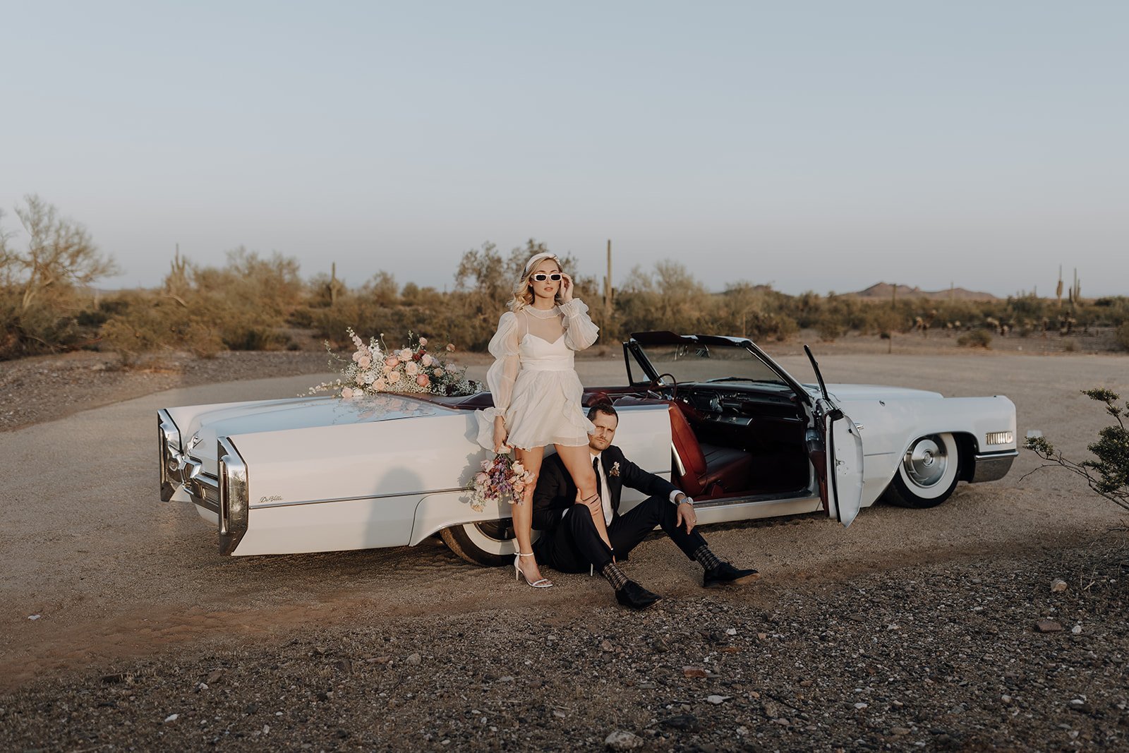 Groom sitting on the ground while bride leans against vintage white convertible