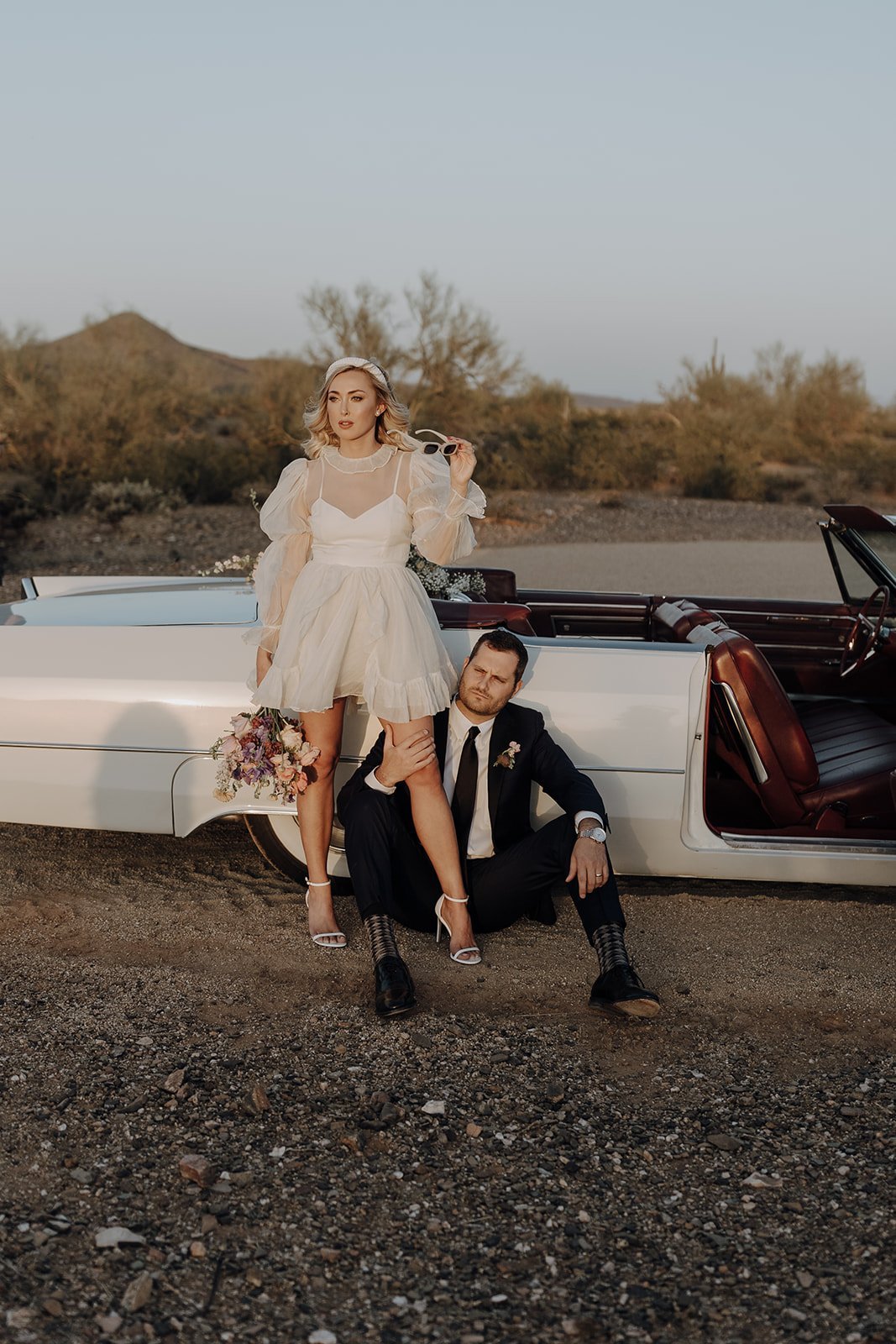 Groom sitting on the ground while bride leans against vintage white convertible