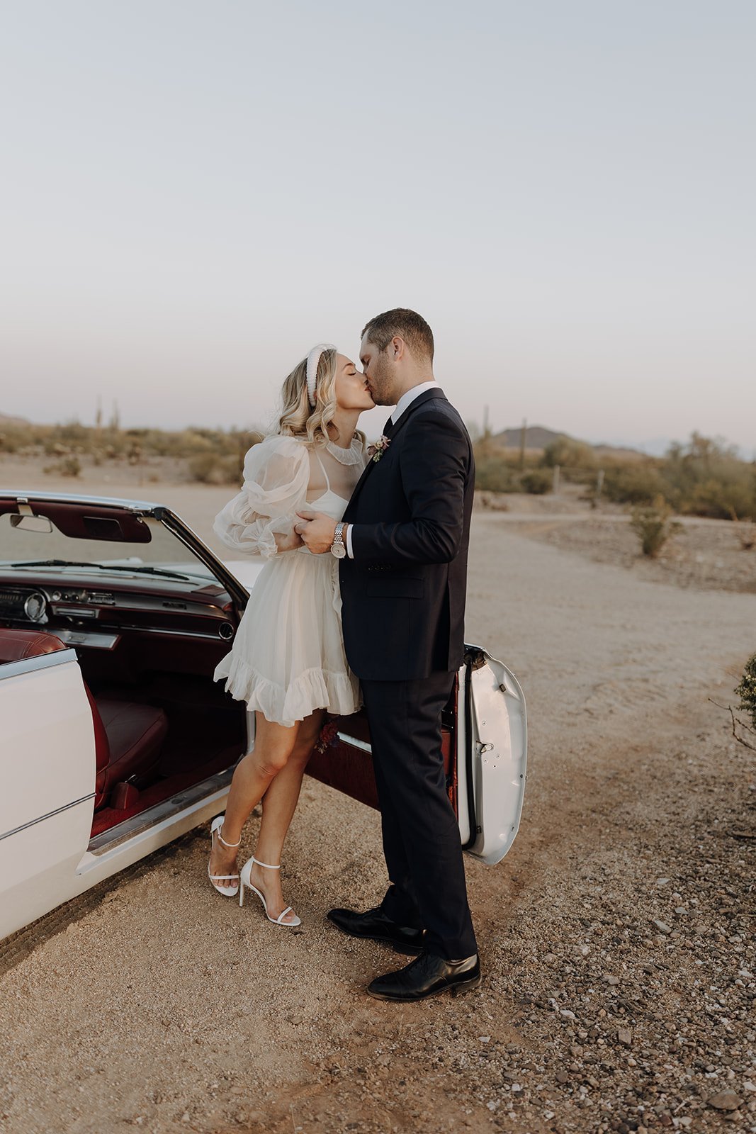 Bride and groom kissing outside of vintage white convertible