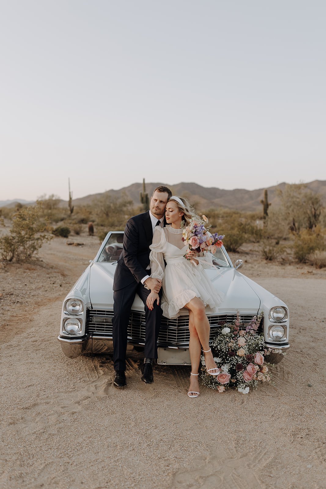 Bride and groom leaning against the front of a vintage white convertible for wedding styled shoot