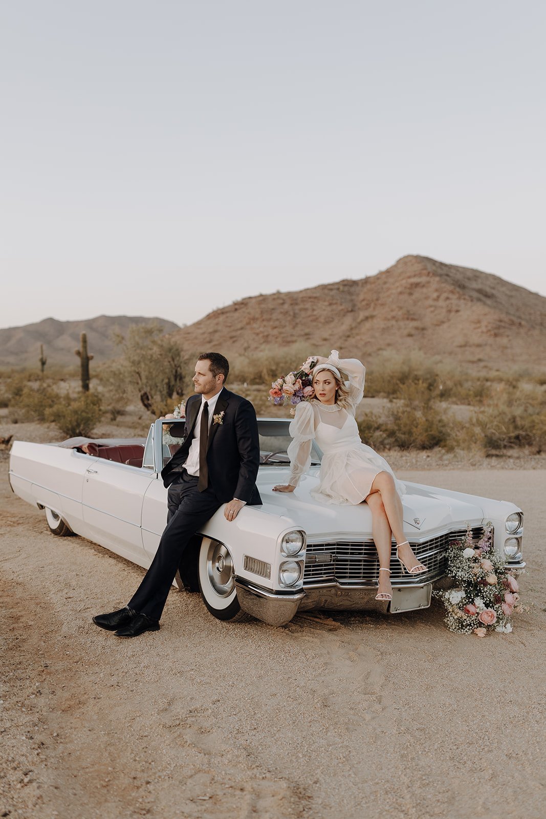 Bride and groom sitting on the hood of a vintage white convertible