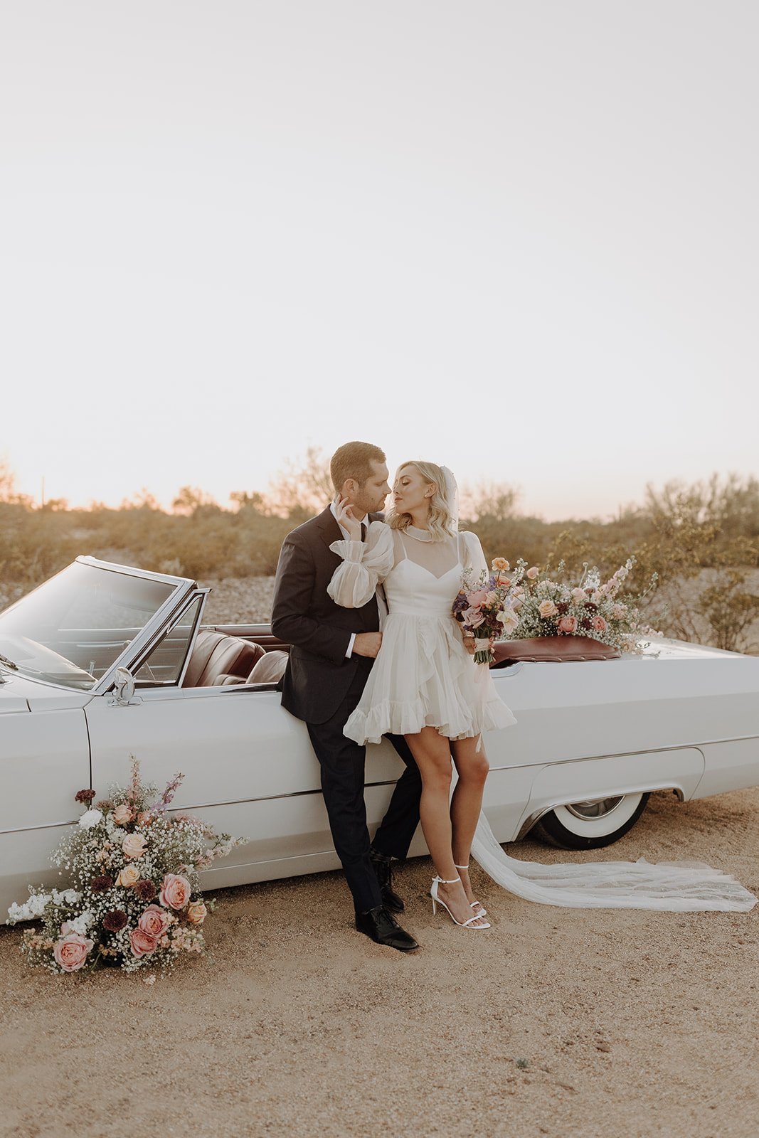 Bride and groom leaning against the side of a vintage white convertible