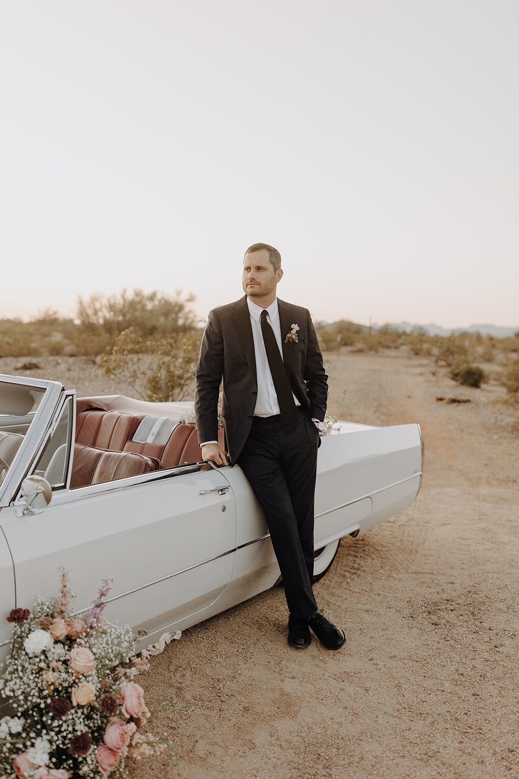 Groom leaning against the side of a vintage white convertible for wedding styled shoot